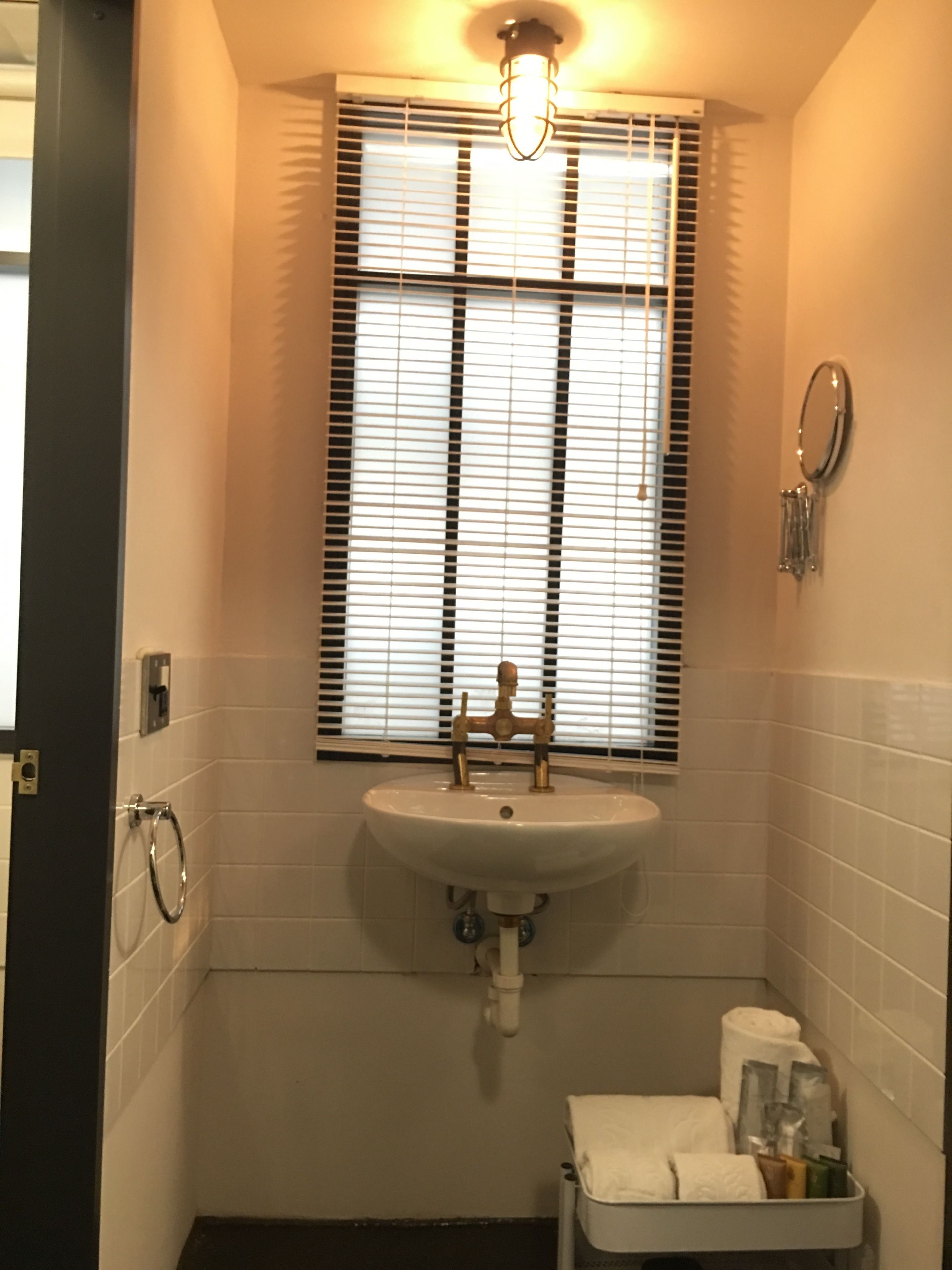 Une salle de bain avec un lavabo blanc, une fenêtre avec des stores, un porte-serviettes et des toilettes avec une poubelle.