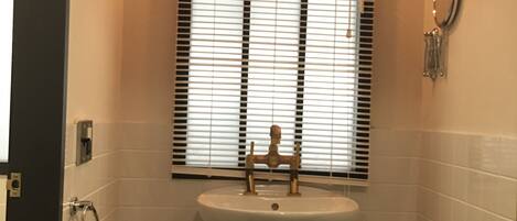 A bathroom with a white sink, a window with blinds, a towel rack, and a toilet with a trash bin.