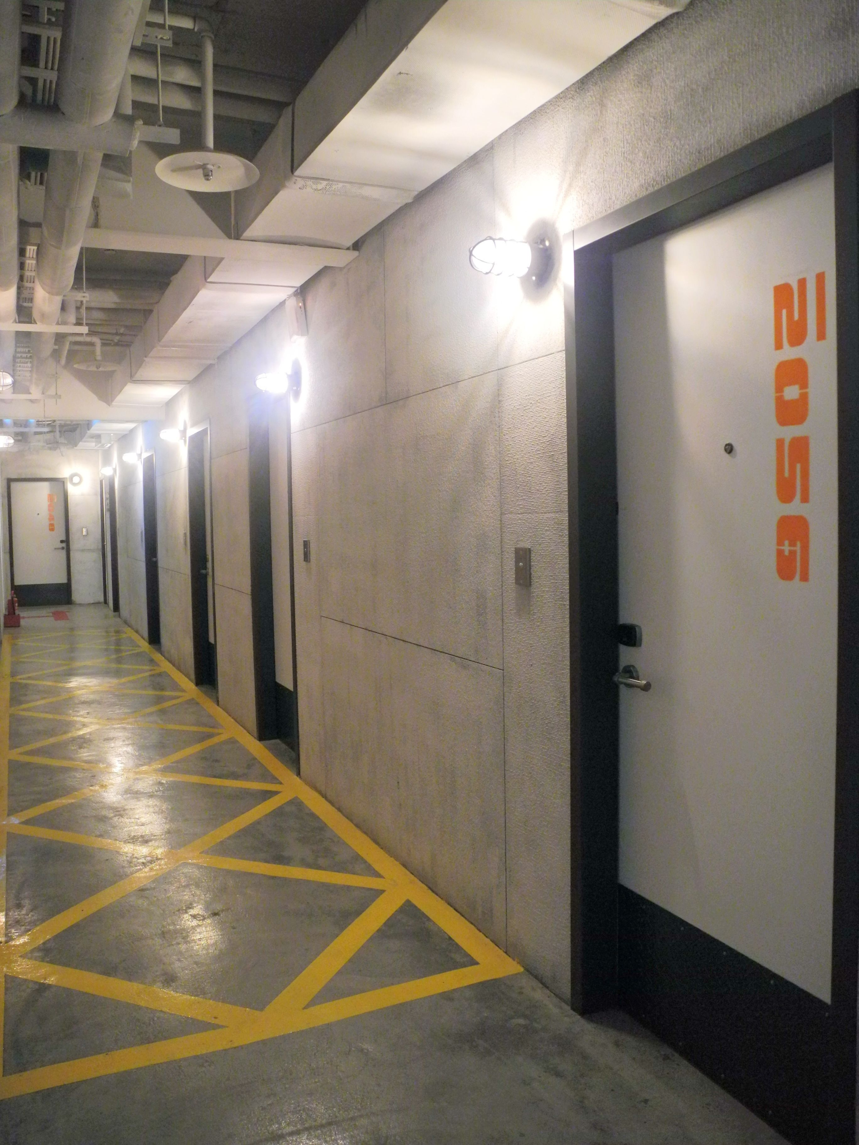 A long, narrow hallway with a yellow geometric pattern on the floor, leading to doors with orange text.