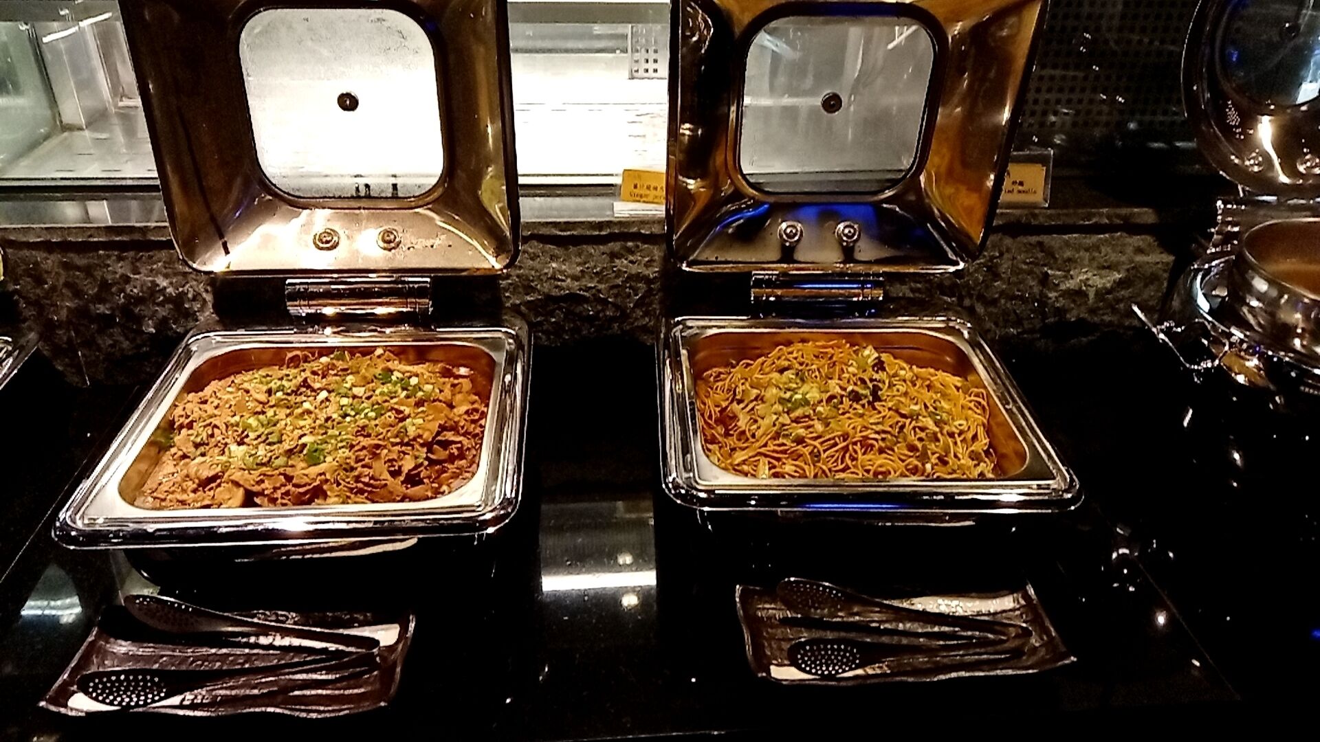Two stainless steel food dispensers with a variety of dishes, including noodles and salads, on a black countertop.