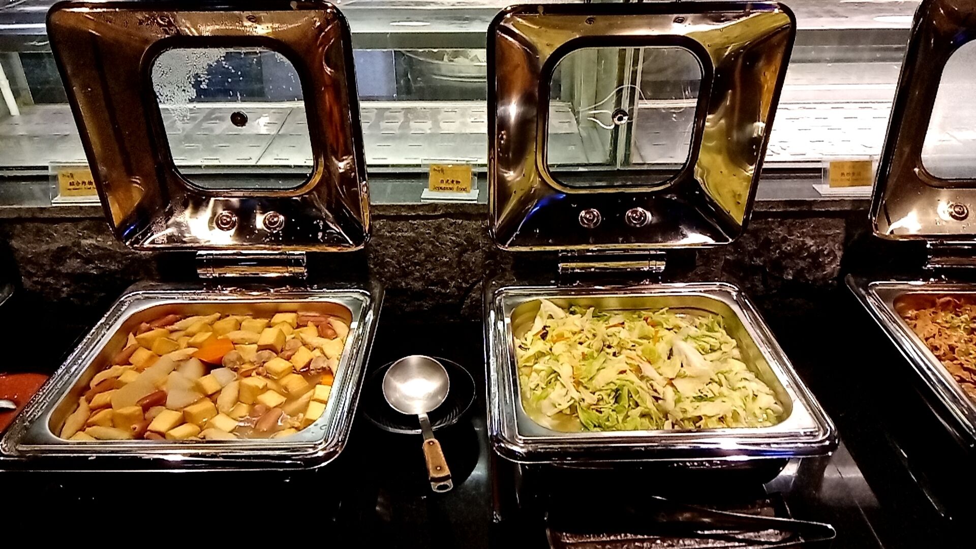 Een buffet met verschillende soorten eten in glazen bakken, waaronder een schotel met blokjes aardappelen en wortels, en een andere met gesneden kool.