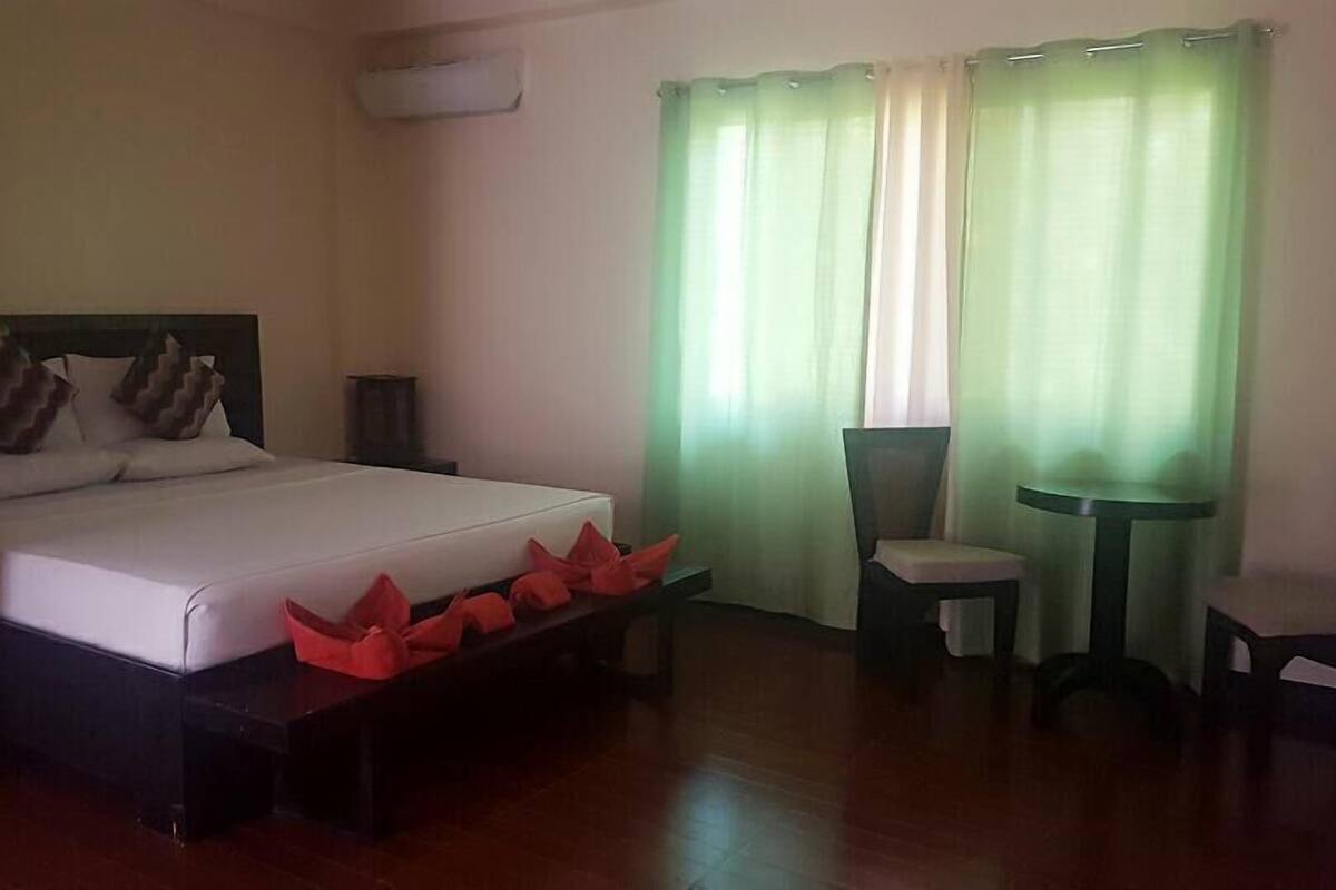 Une chambre d’hôtel avec un lit, une petite table, deux chaises et une fenêtre avec des rideaux verts.