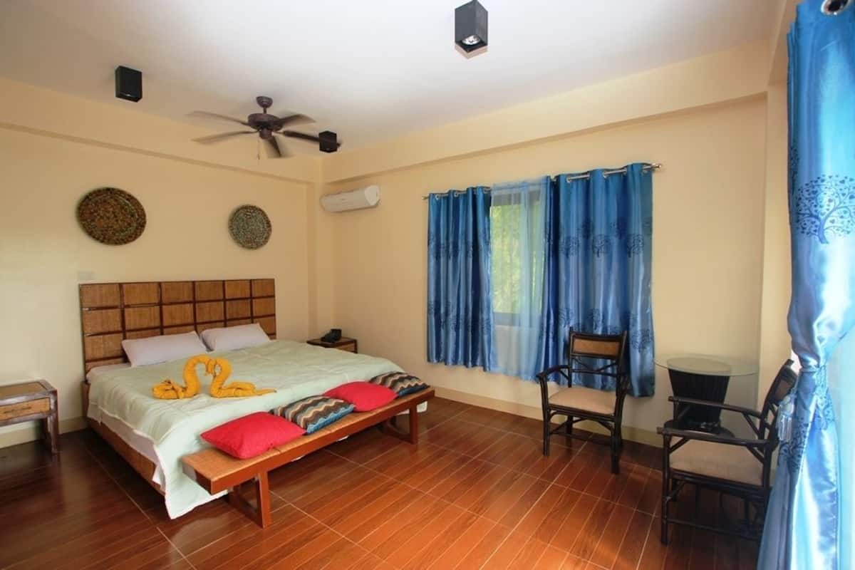 Une chambre à coucher avec un lit, un ventilateur de plafond, une fenêtre avec des rideaux bleus et une petite table avec des chaises.