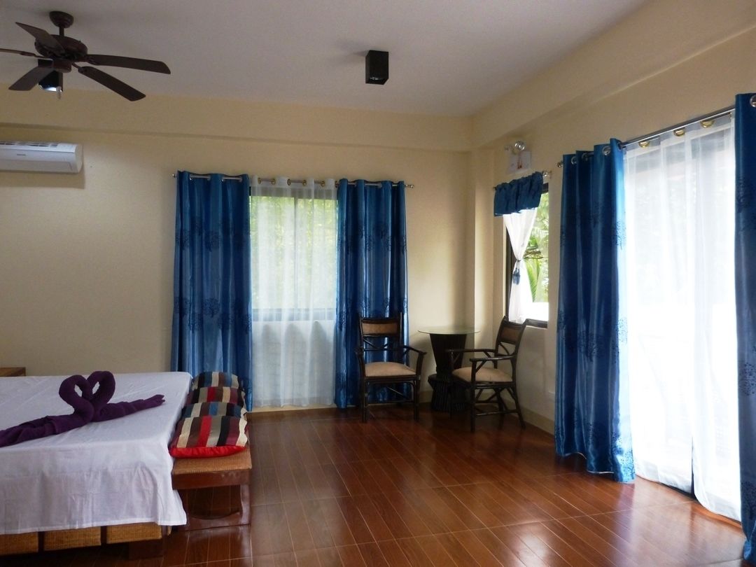 Una habitación de hotel con una cama, un ventilador de techo, una mesa con sillas y cortinas azules.