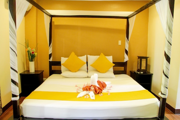 Habitación de hotel con una cama grande, dos almohadas, una mesita de noche, un jarrón con flores y una colcha amarilla.