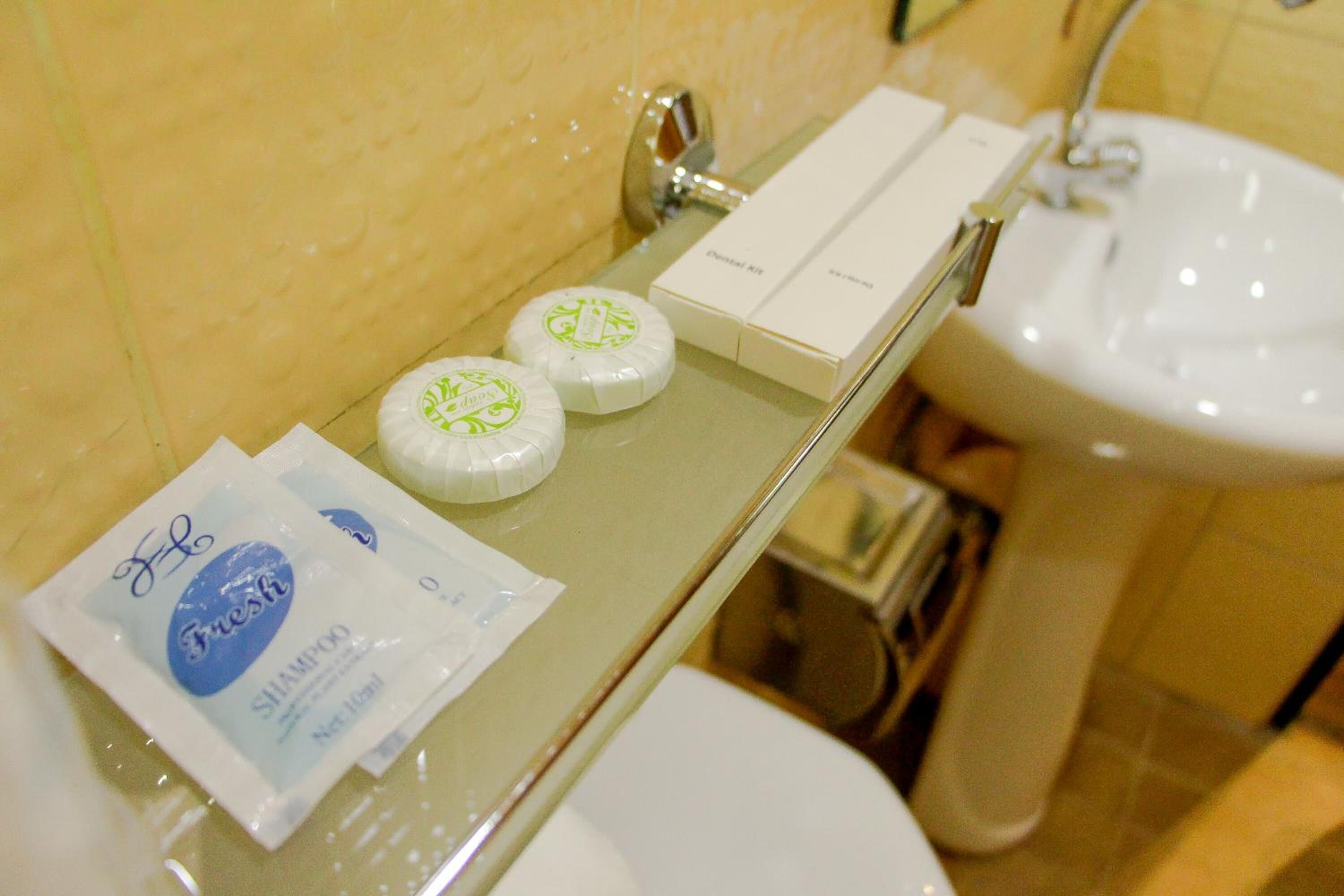 Casa de banho com divisória de vidro, lavatório branco e produtos de higiene, incluindo um frasco novo de champô e duas barras de sabão.
