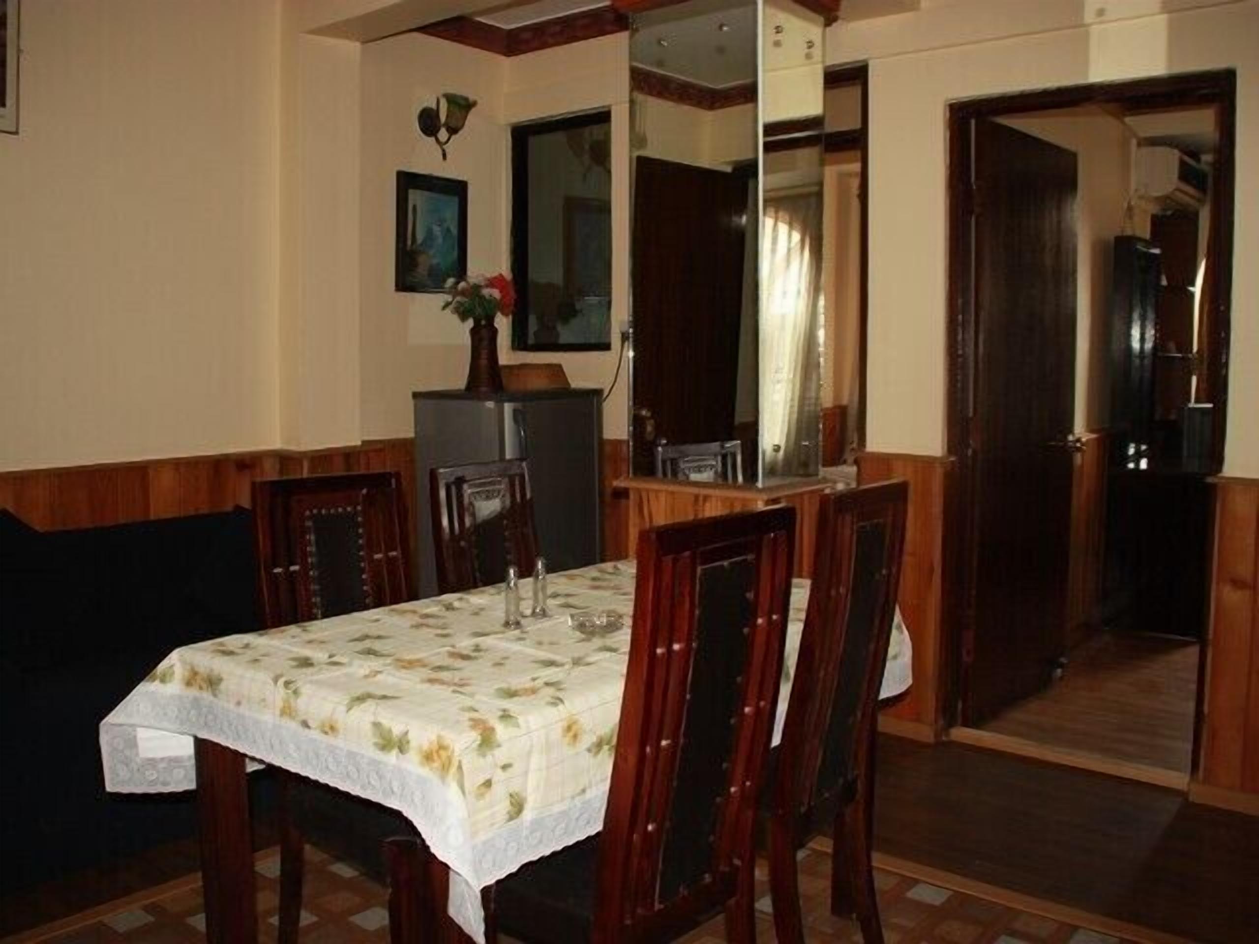 Een eetruimte met een tafel voor vier personen, omringd door houten stoelen. Een koelkast staat tegen de muur, en een spiegel weerspiegelt een deel van de kamer.