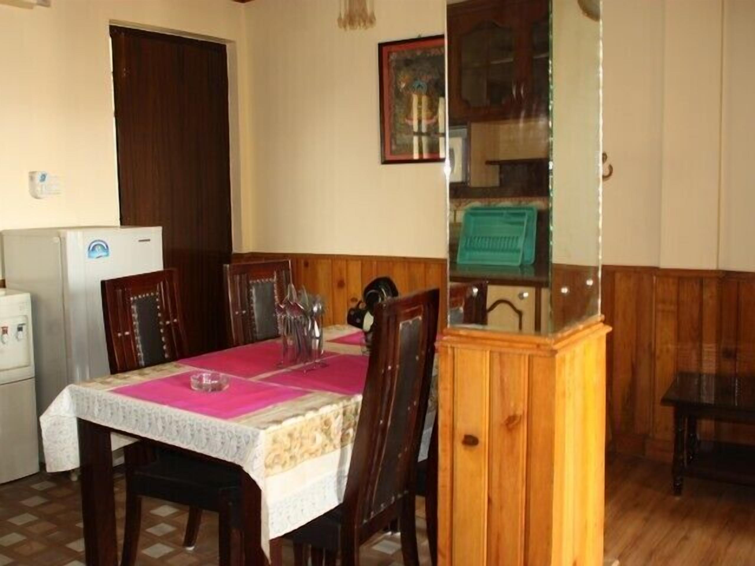 Een eetgedeelte met een tafel, bedekt met een roze tafelkleed, houten stoelen, een houten kast en een koelkast.