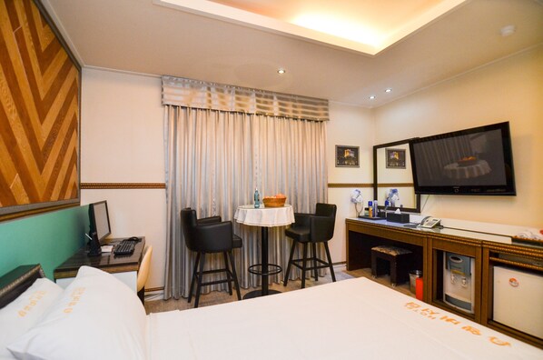 Una habitación de hotel con una cama, un escritorio con una computadora, dos sillas, una mesita con un tazón, un televisor y un minibar.