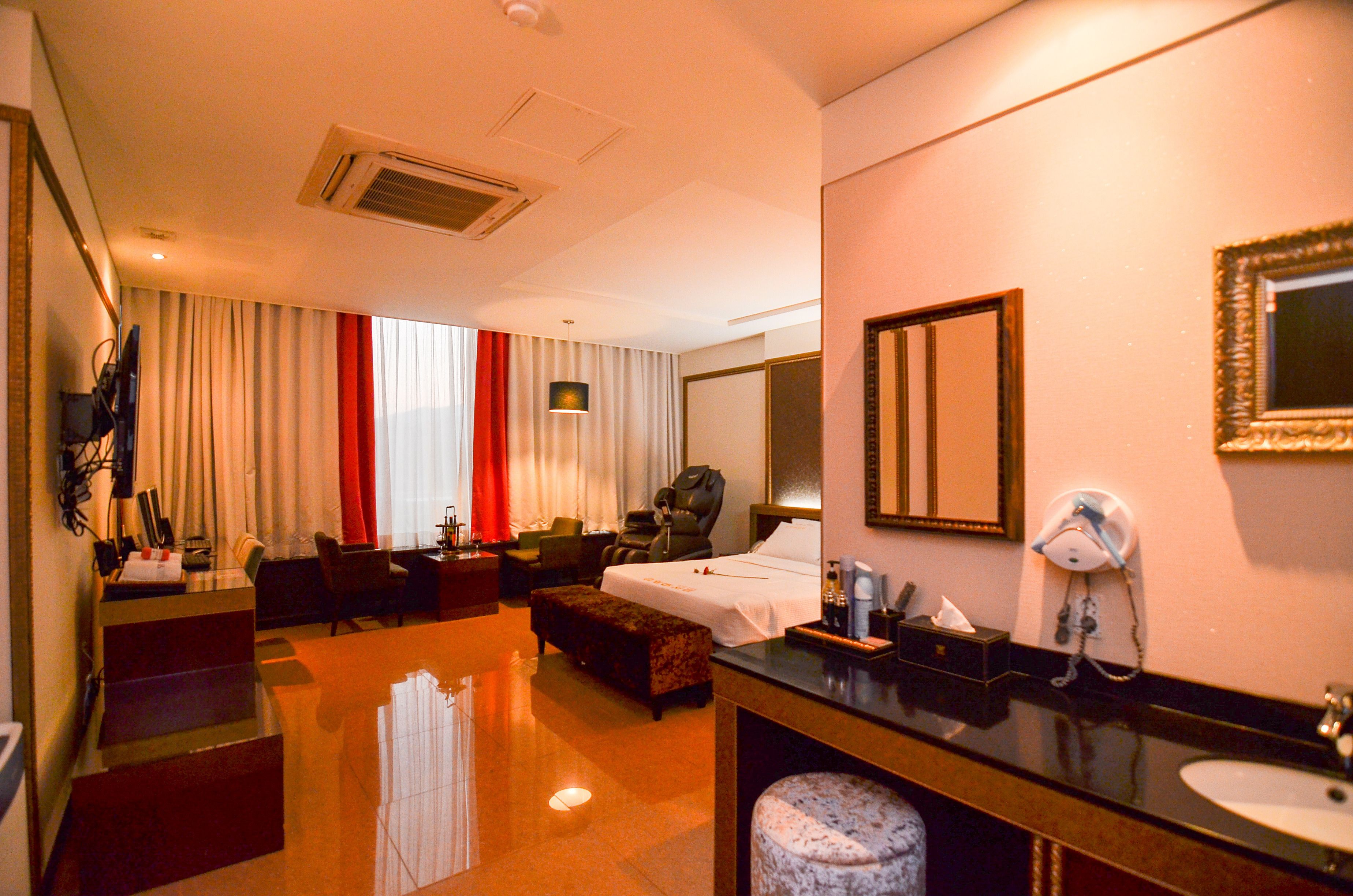 A hotel room with a bed, a desk, a chair, a sofa, a bathroom with a sink and mirror, and a TV.