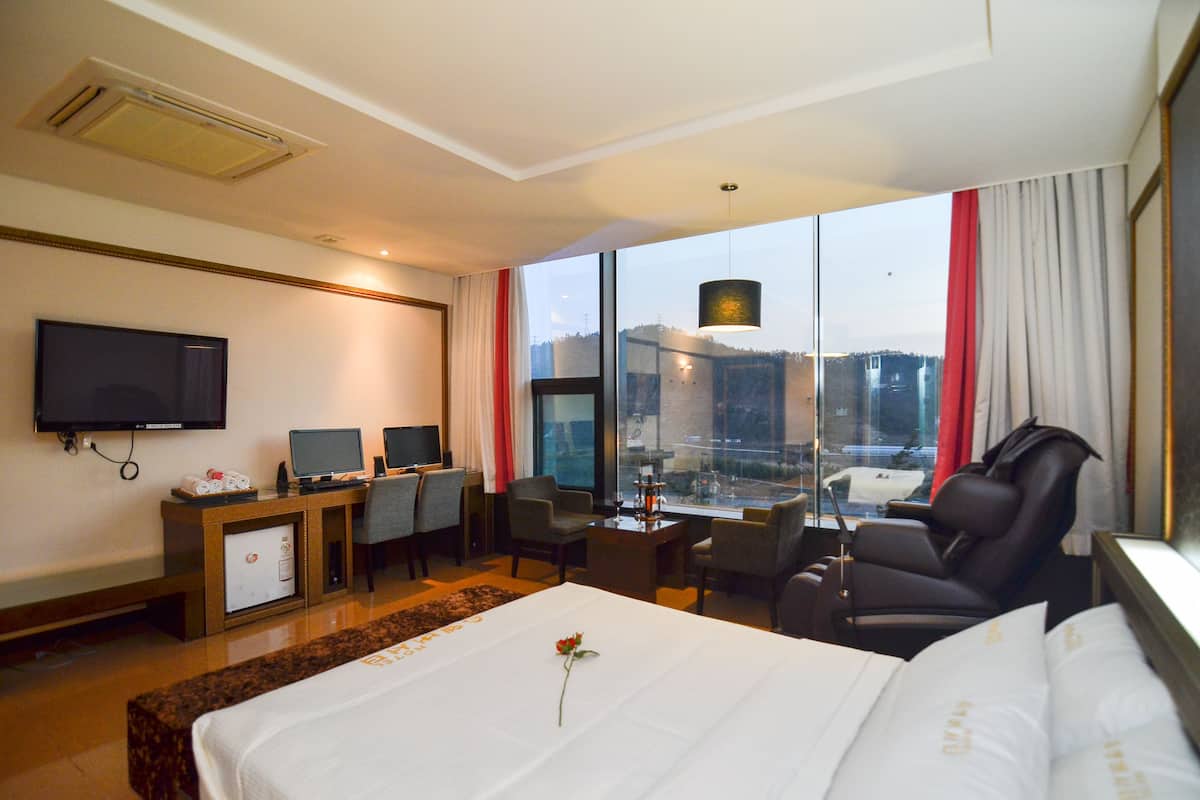 A hotel room with a large bed, a desk with two monitors, a TV, and a view of the city through large windows.