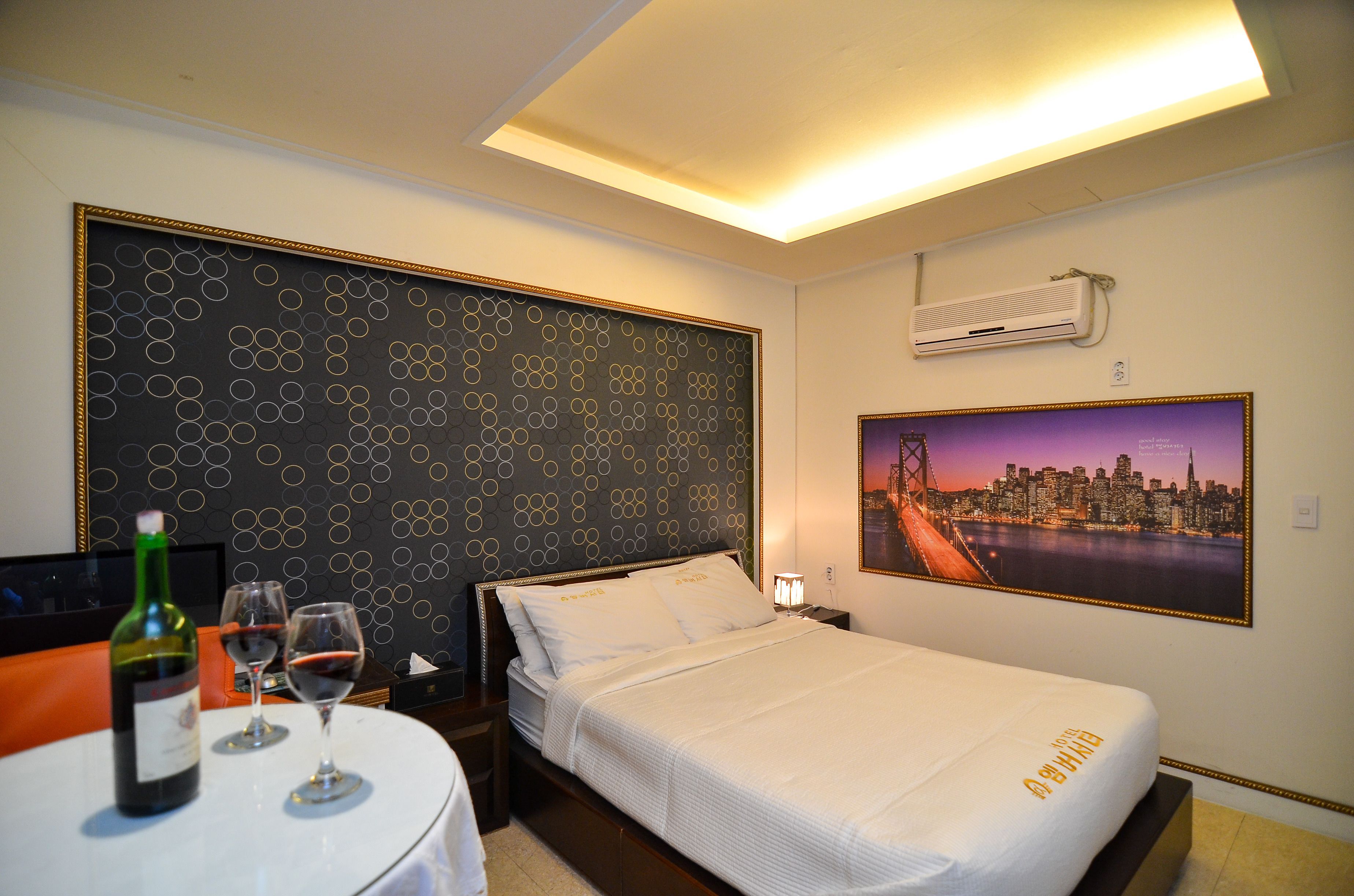 A hotel room with a bed, a table with a bottle and glasses, a cityscape painting, and a patterned headboard.