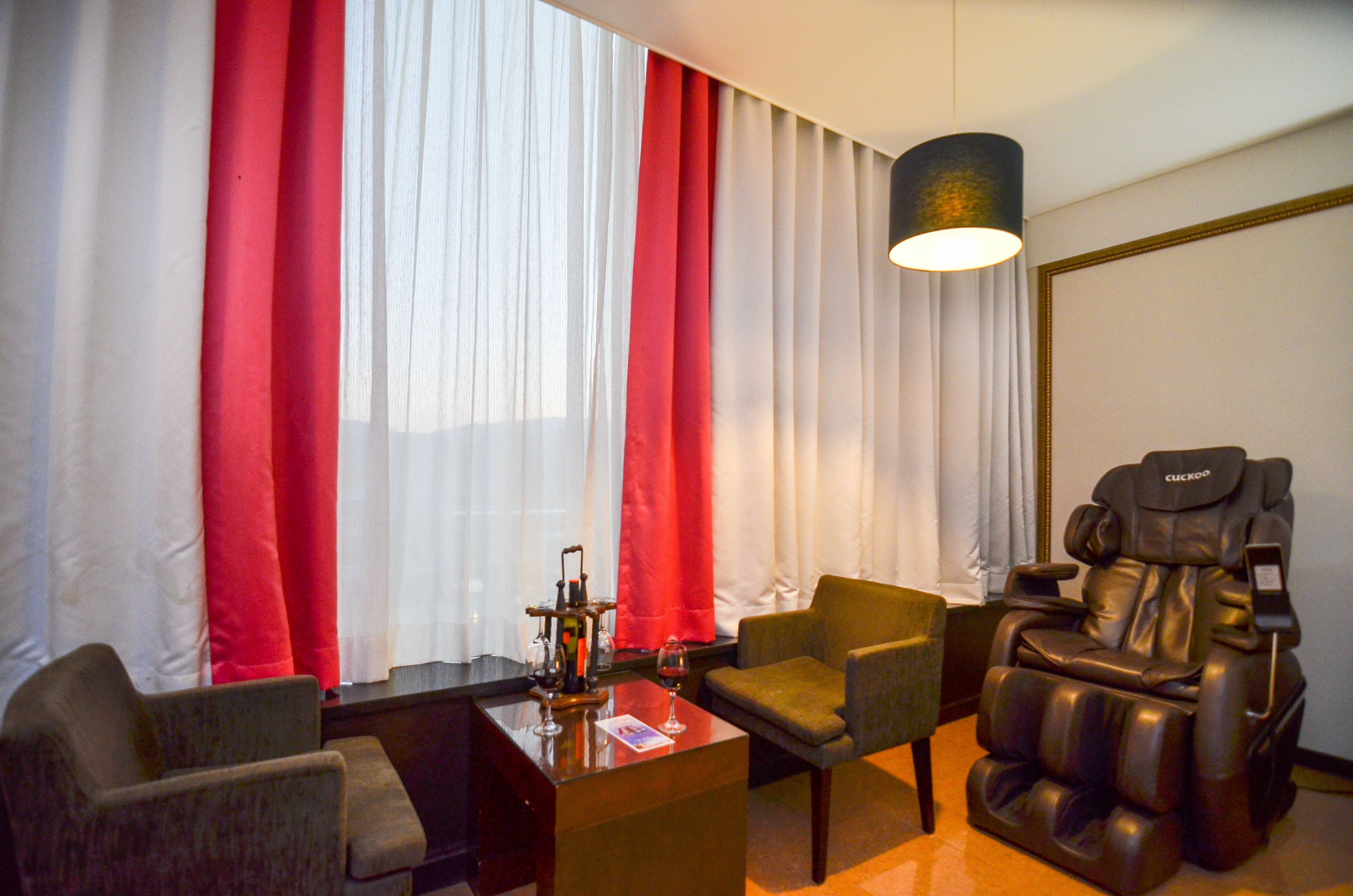 A room with a massage chair, two armchairs, a coffee table with a wine glass, and red and white curtains.