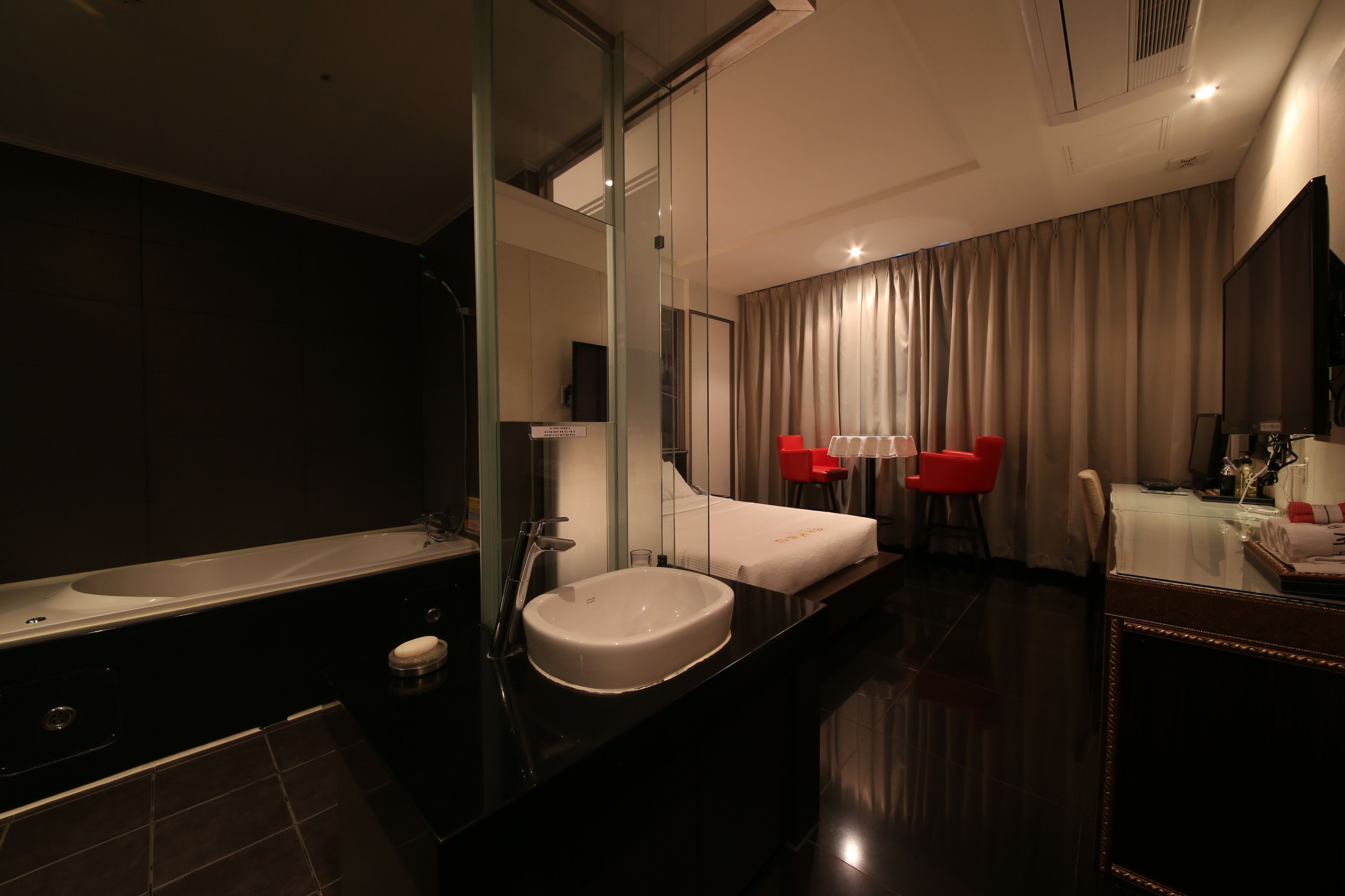 A modern hotel bathroom with a large mirror, a bathtub, a sink, and two red chairs.