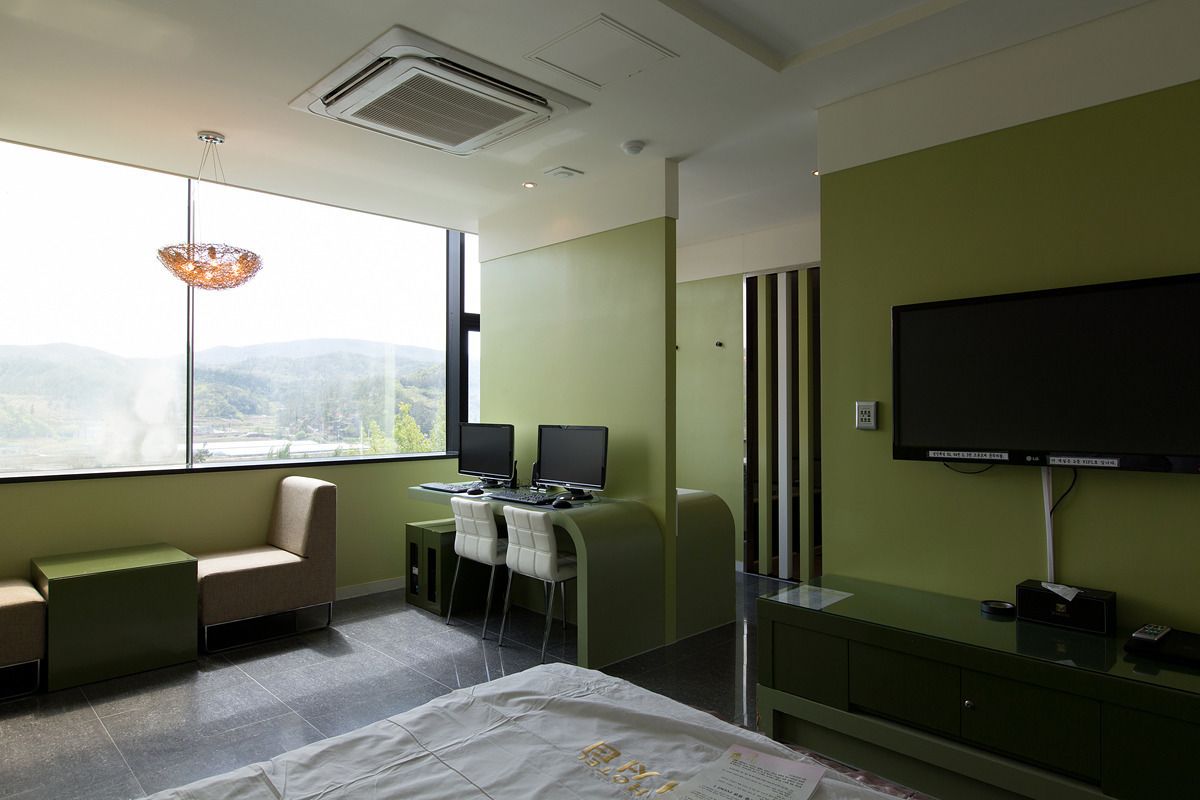 Ein modernes Hotelzimmer mit großem Fenster, zwei Schreibtischen mit Computern, einem Bett und einem Flachbildfernseher.