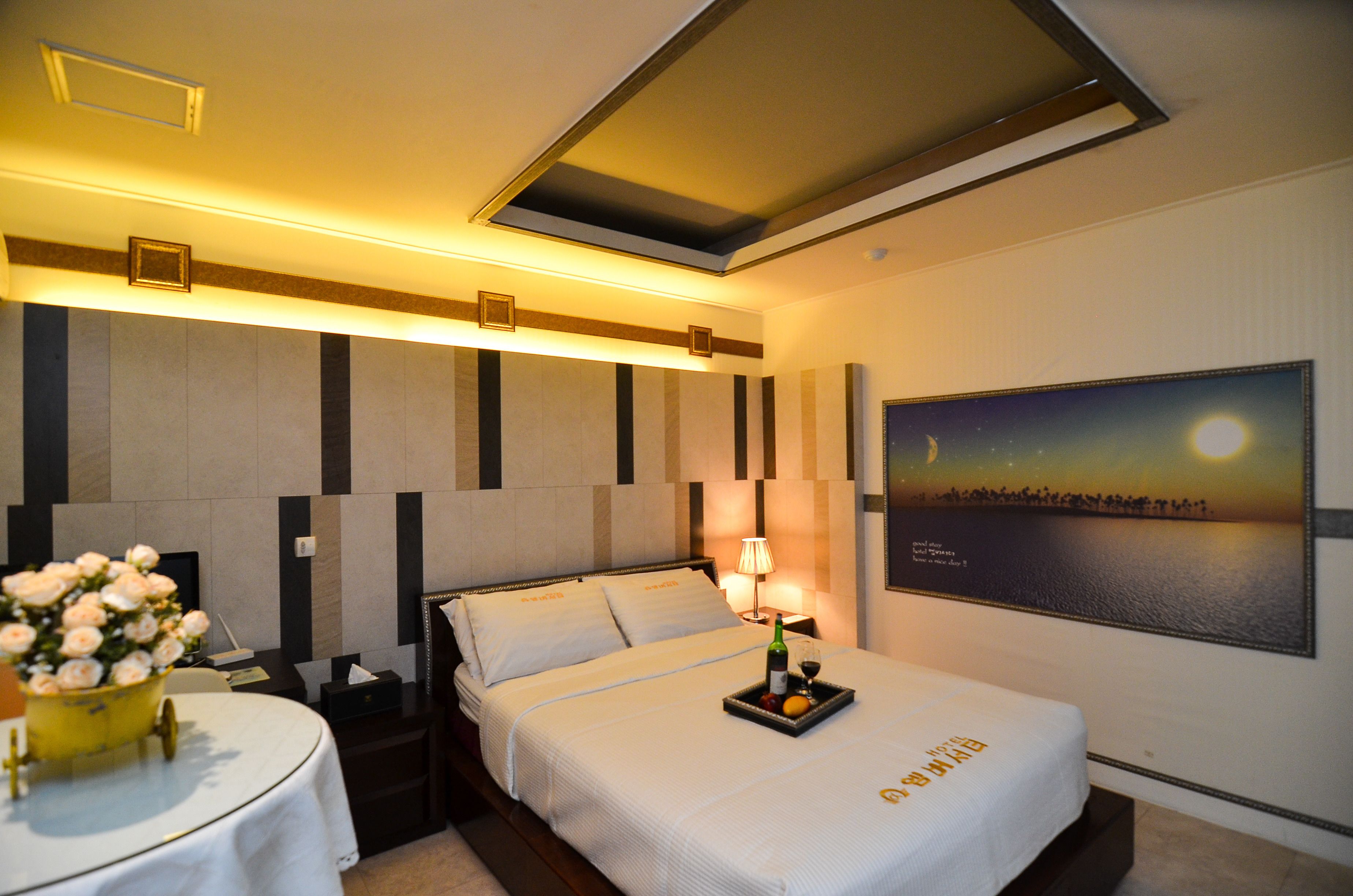 Una habitación de hotel con cama, mesita de noche, lámpara y un cuadro de un atardecer.