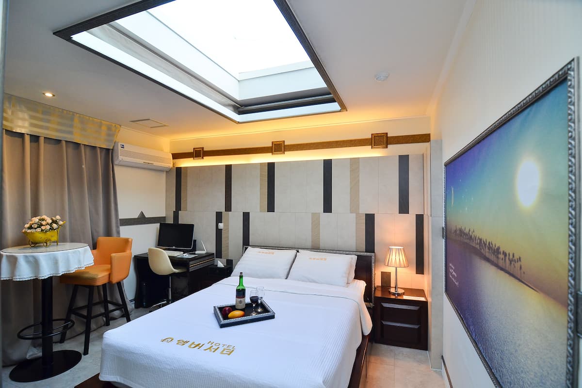 A hotel room with a bed, a desk, a chair, a table with a tray of fruit and a bottle, and a painting on the wall.