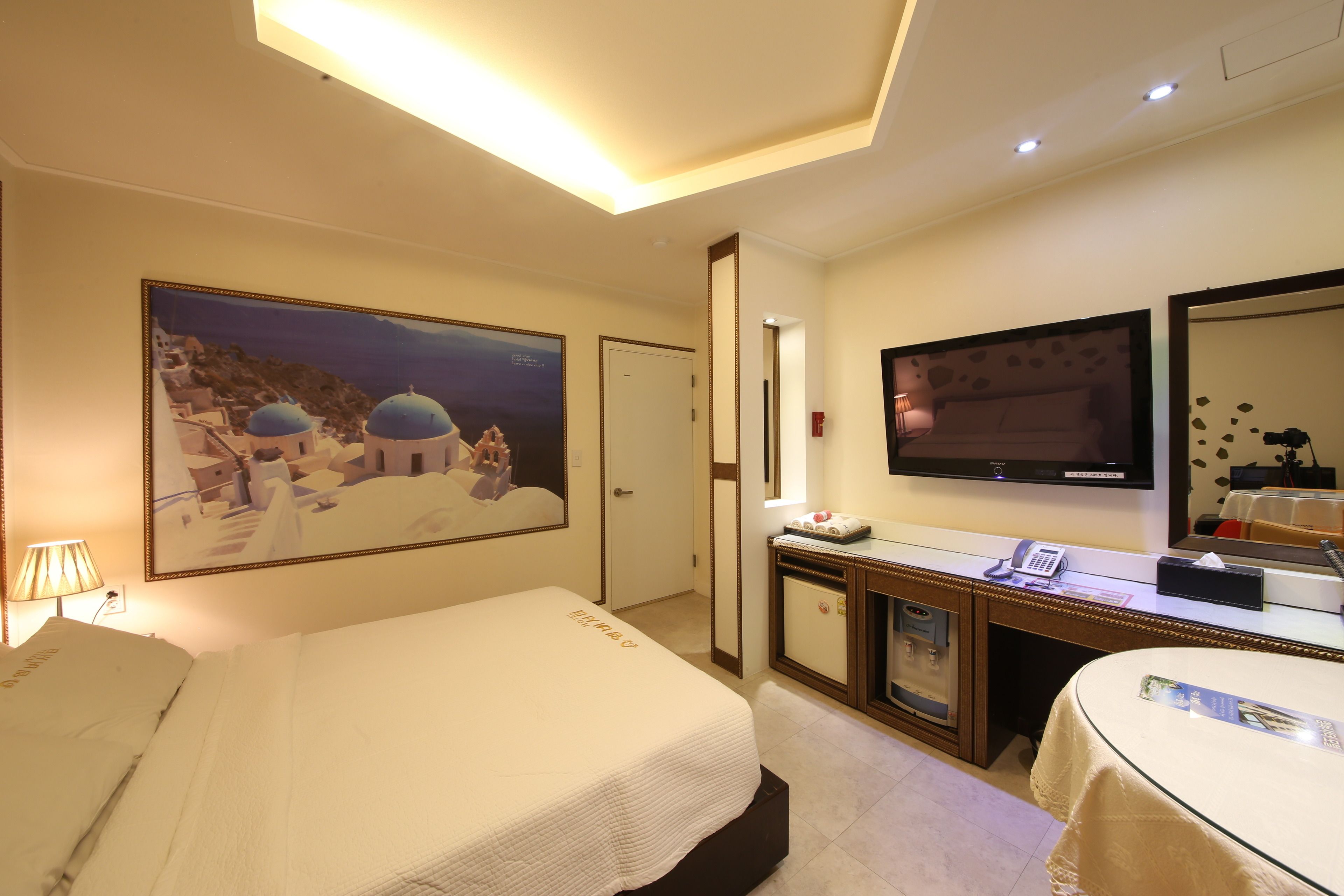 A hotel room with a bed, a desk with a TV, a mirror, and a painting of a coastal scene.