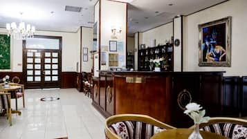 A hotel reception area with a reception desk, chairs, a chandelier, framed artwork, and a door with frosted glass panels.