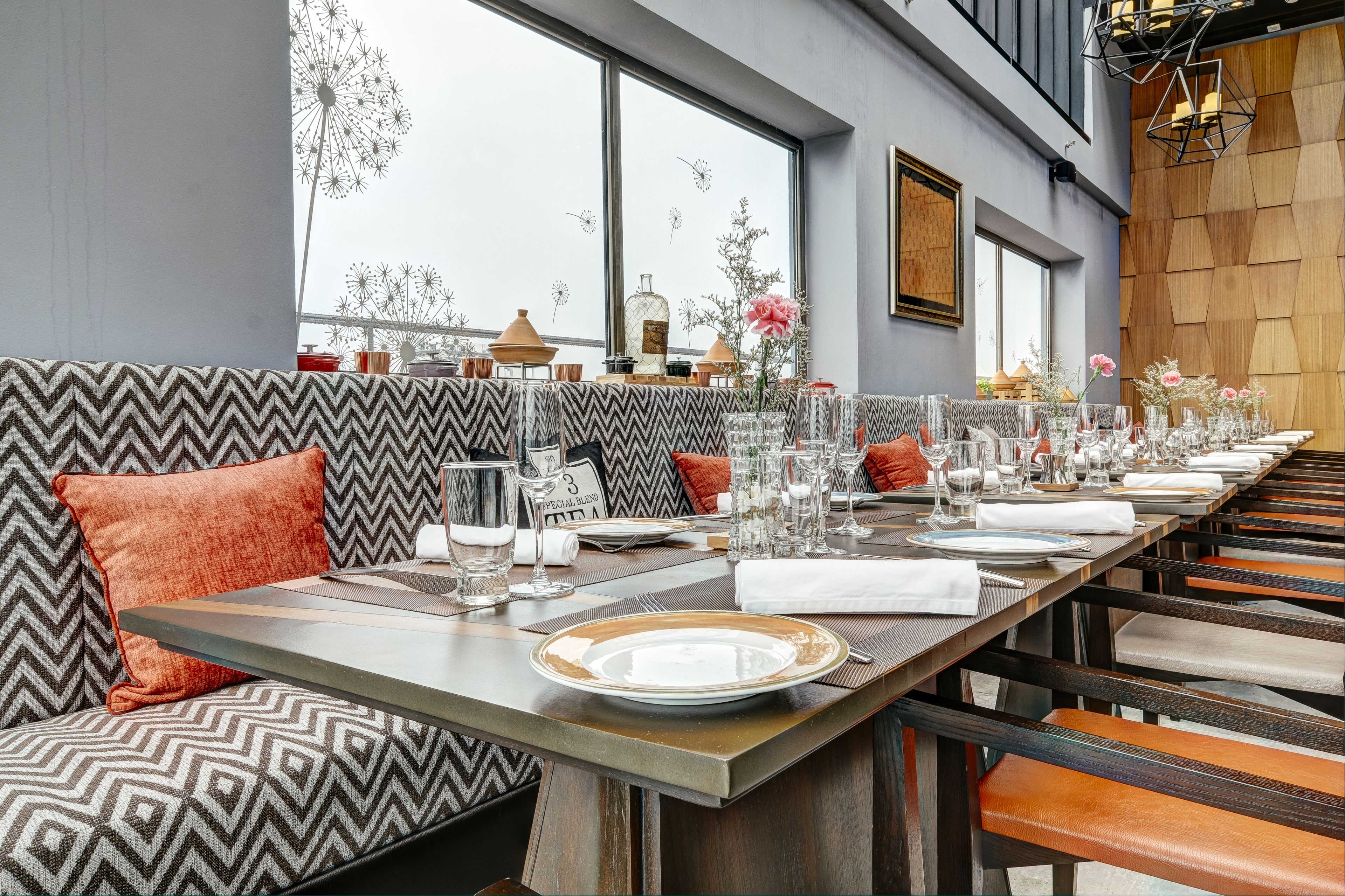 A restaurant with a long dining table set for a meal, a patterned bench, and a view of dandelions through large windows.
