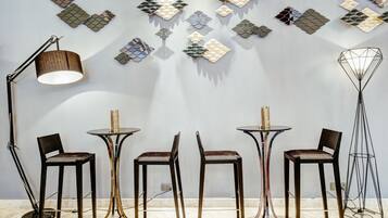 Un interior moderno con un arte mural geométrico, una lámpara de pie y otra lámpara de pie alta y estilizada. Hay cuatro sillas y dos mesas.