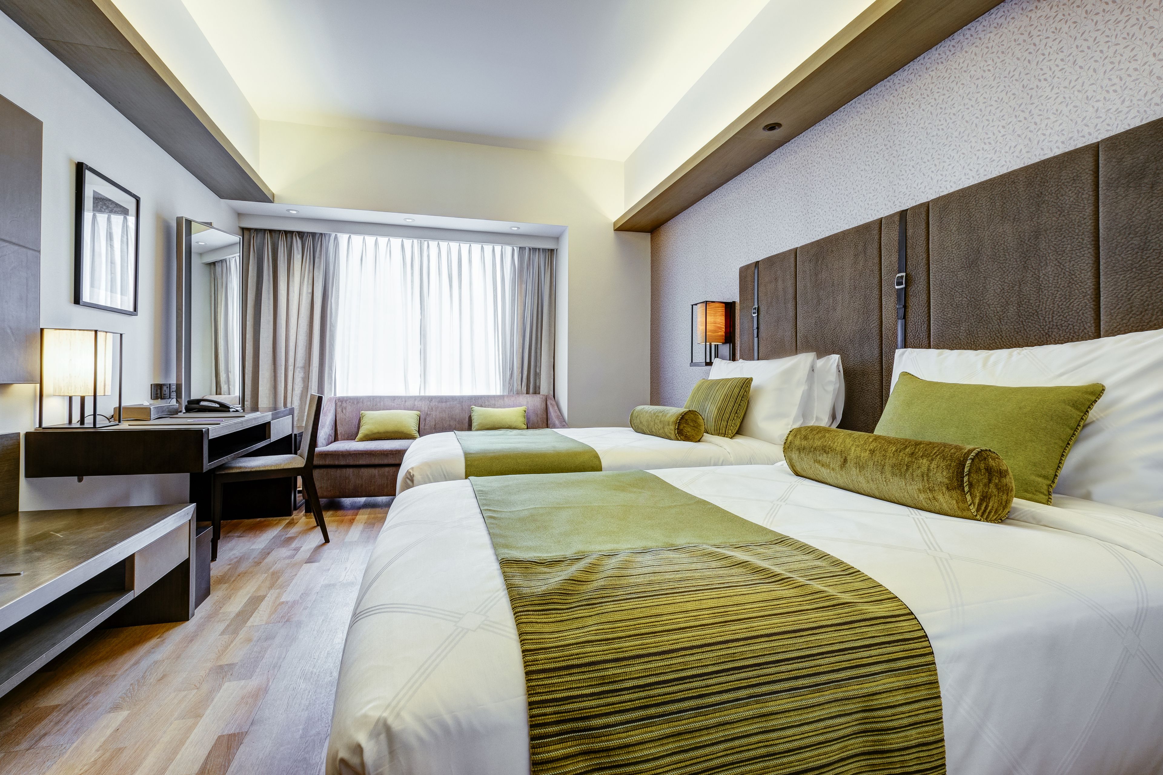 A hotel room with a large bed, a desk, and a chair. There is a window with curtains, a lamp, and a mirror on the wall.
