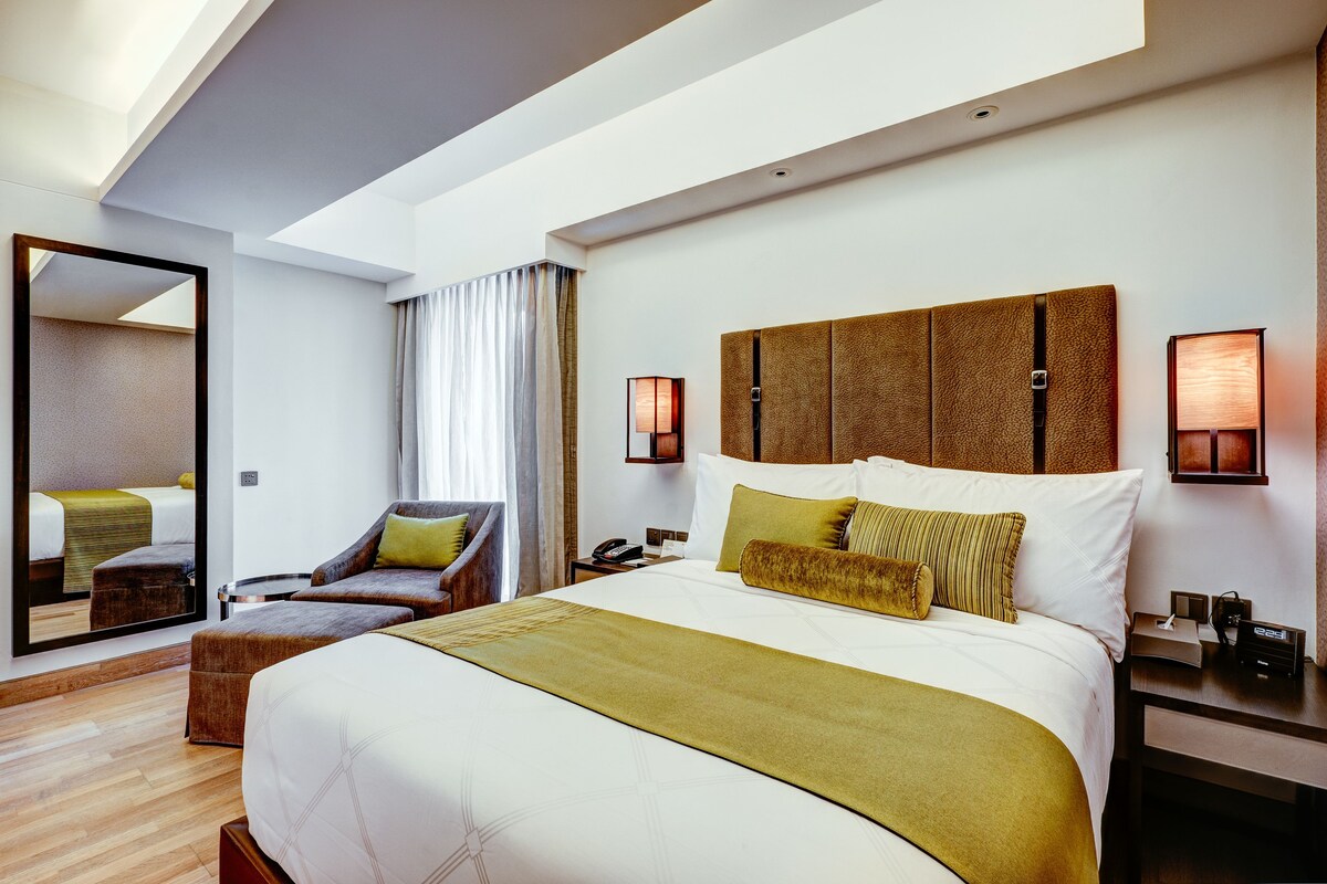 Ein modernes Hotelzimmer mit einem großen Bett, einer Sitzecke mit Sessel und Hocker, einem Schreibtisch mit Lampe und einem großen Spiegel.