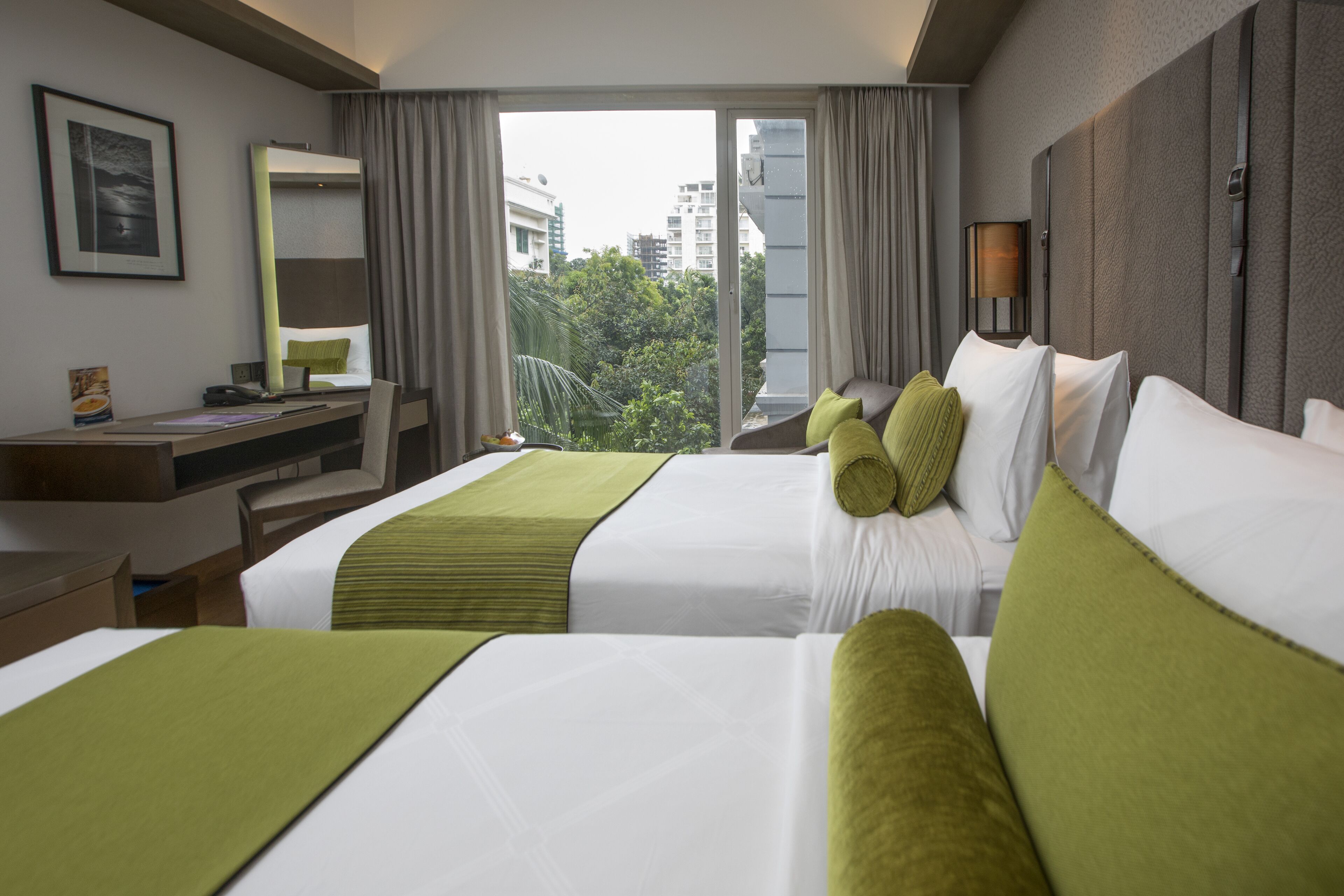 Habitación de hotel con dos camas, un escritorio, una silla, un ventanal y vistas a edificios y árboles.