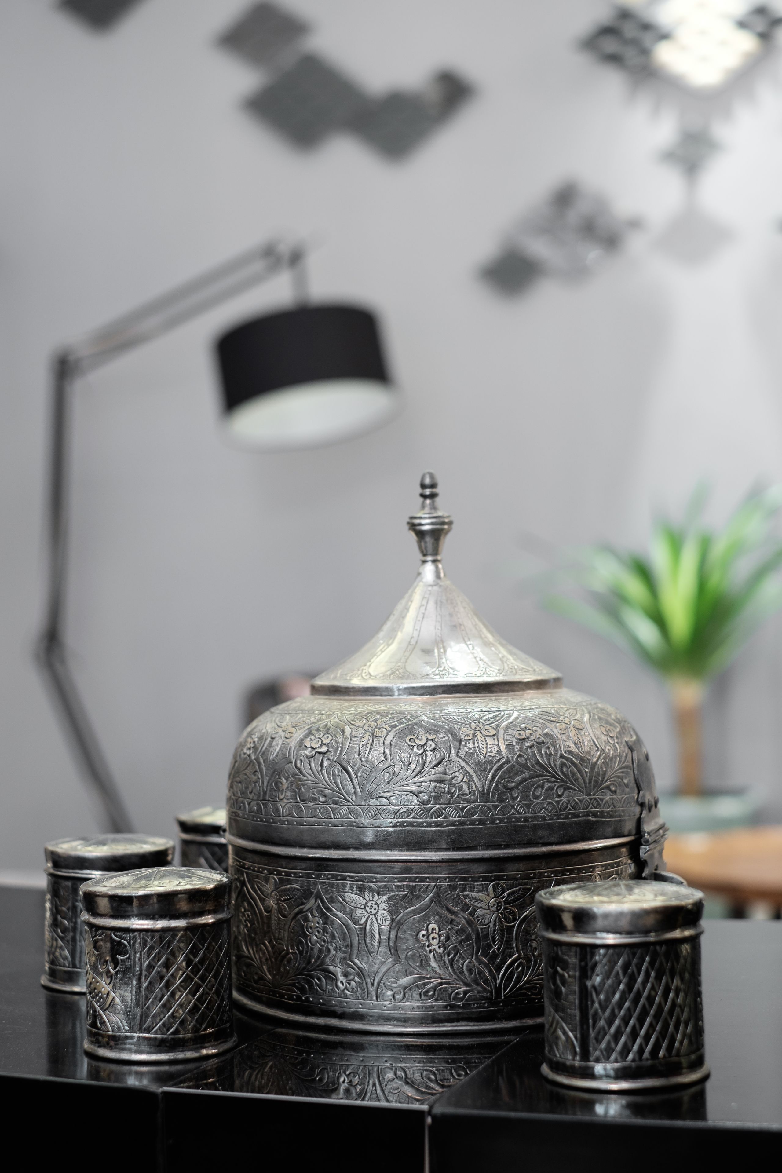 A decorative metal container with intricate designs, accompanied by smaller containers and a lamp in a modern setting.