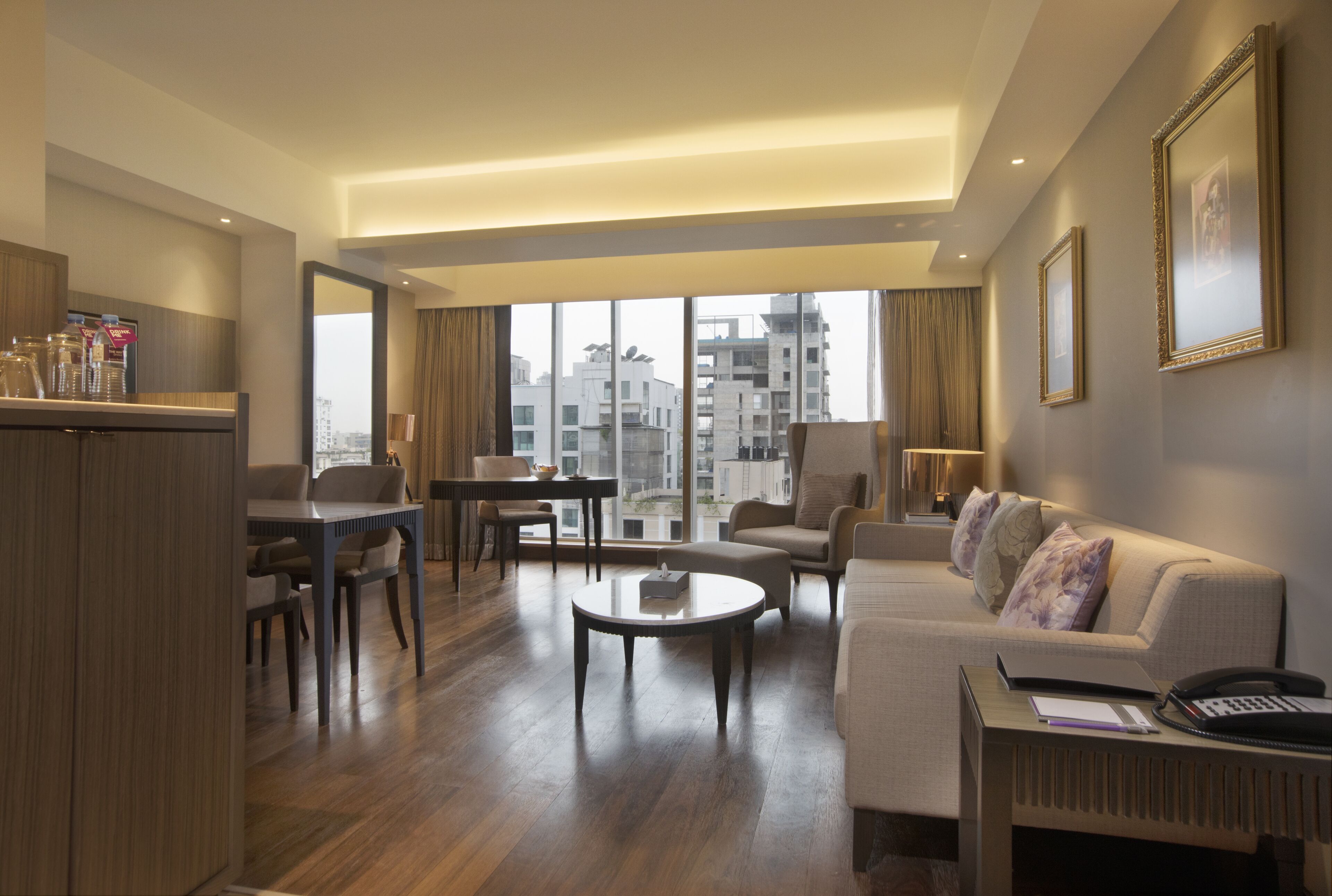 A modern hotel room with a dining area, a sofa, a coffee table, and a desk with a lamp.