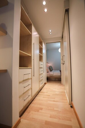 Un couloir étroit, doté d’un plancher en bois, mène à une chambre à coucher qui contient un lit et une table de chevet.