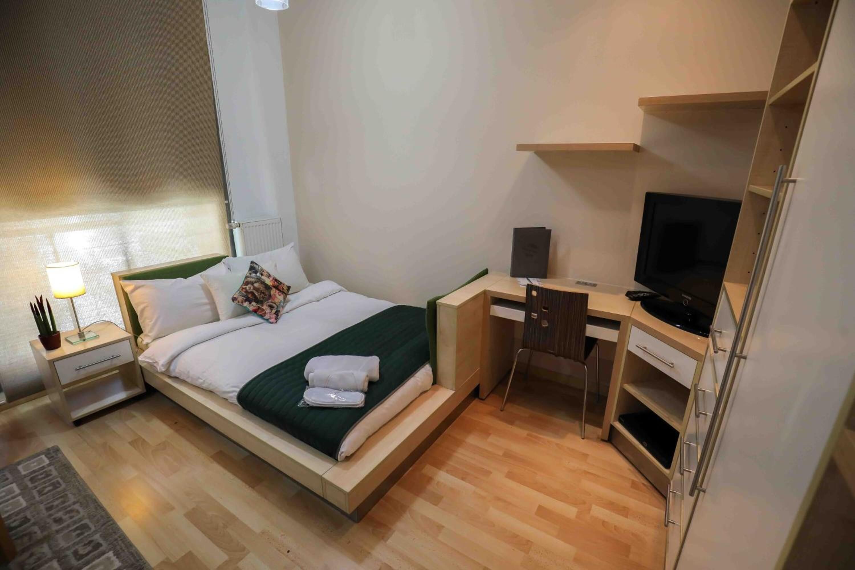 A hotel room with a bed, a desk, a chair, a TV, and a nightstand with a lamp.