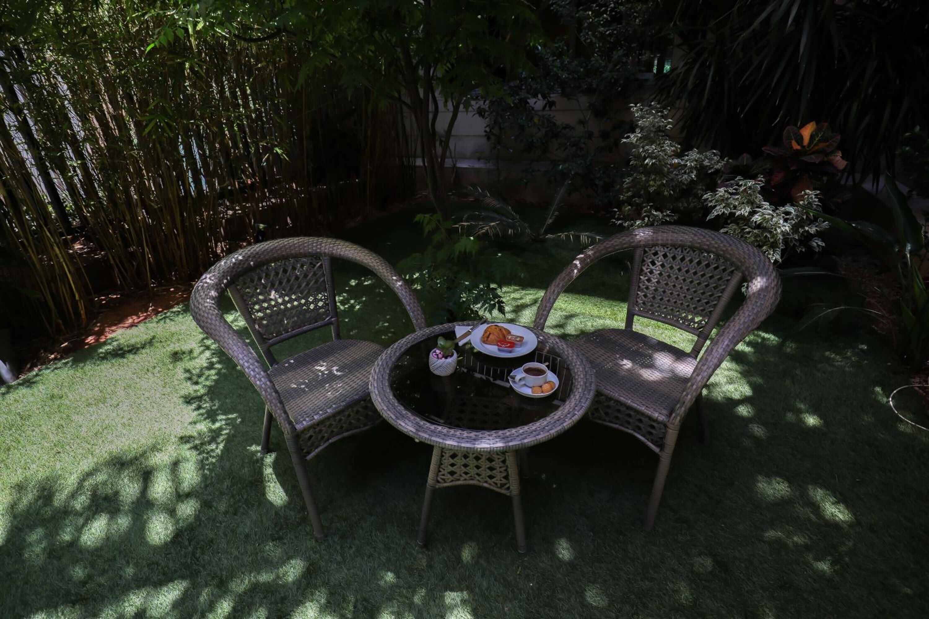 Un pequeño espacio exterior con sillas de mimbre y una mesa redonda, rodeado de abundante vegetación y bambú.