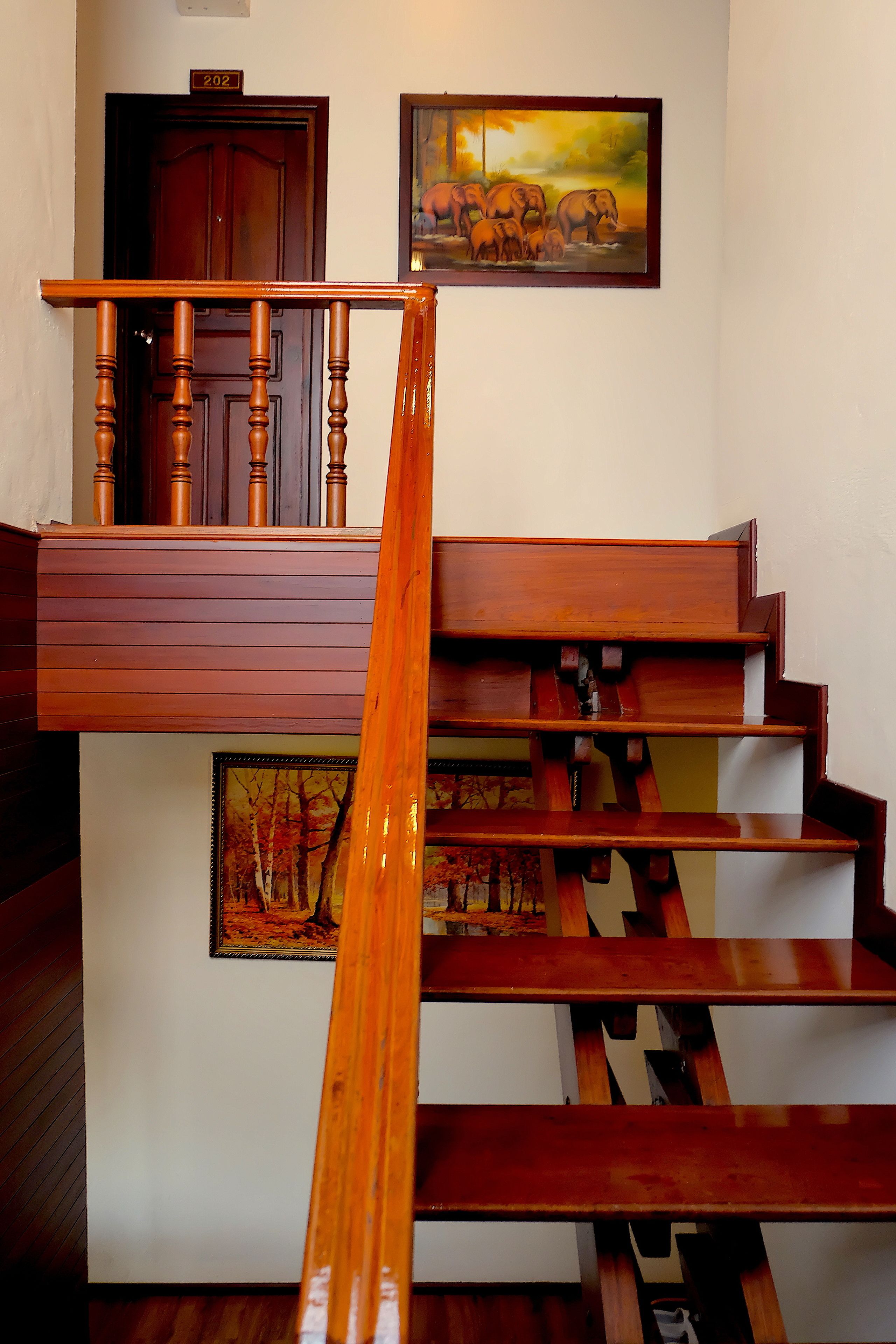 Un escalier en bois avec une rampe, une porte portant le numéro 202 et deux tableaux encadrés accrochés au mur.