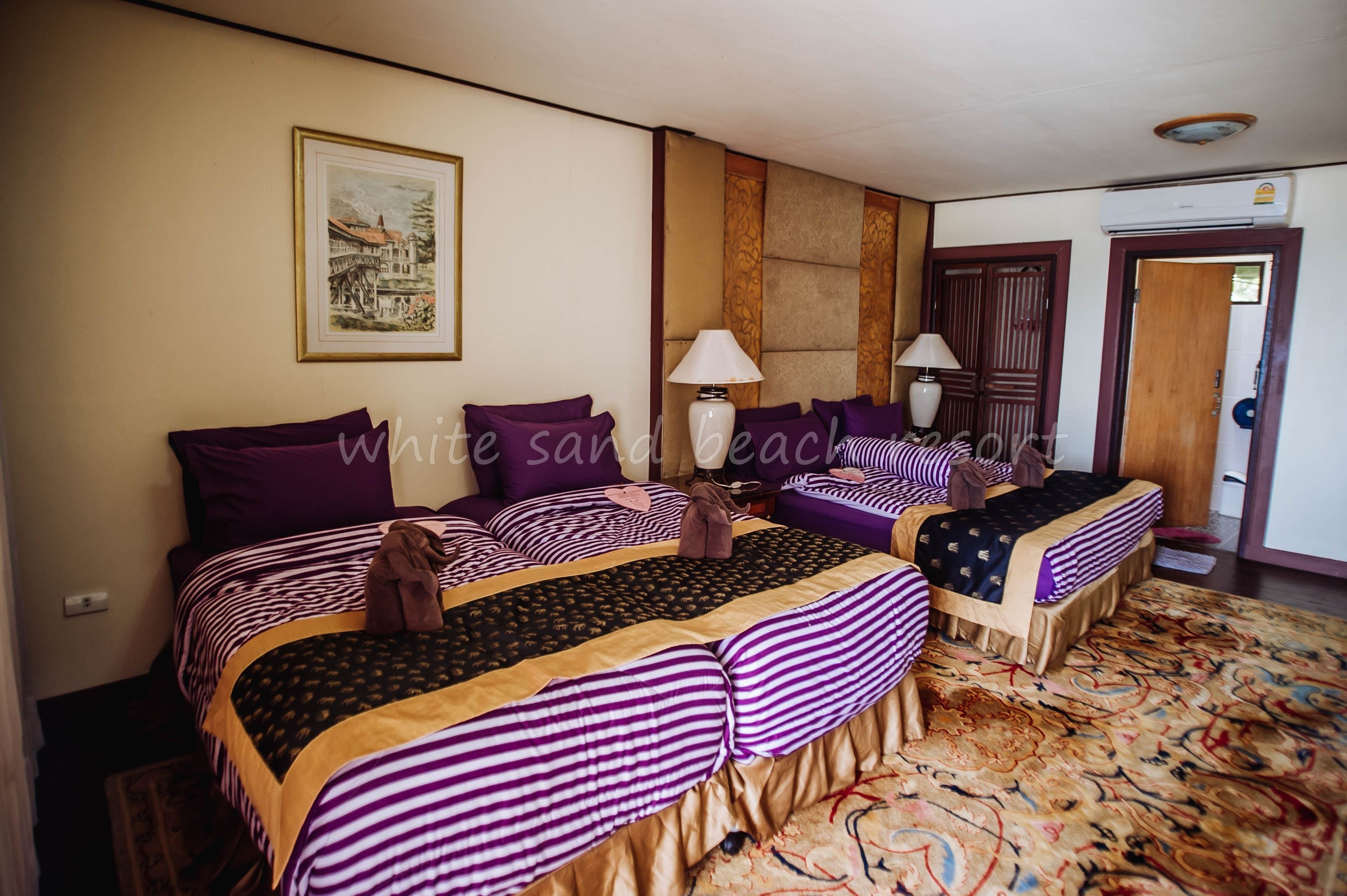 Habitación de hotel con dos camas, cada una con ropa de cama morada y dorada, una alfombra grande estampada y un cuadro en la pared.