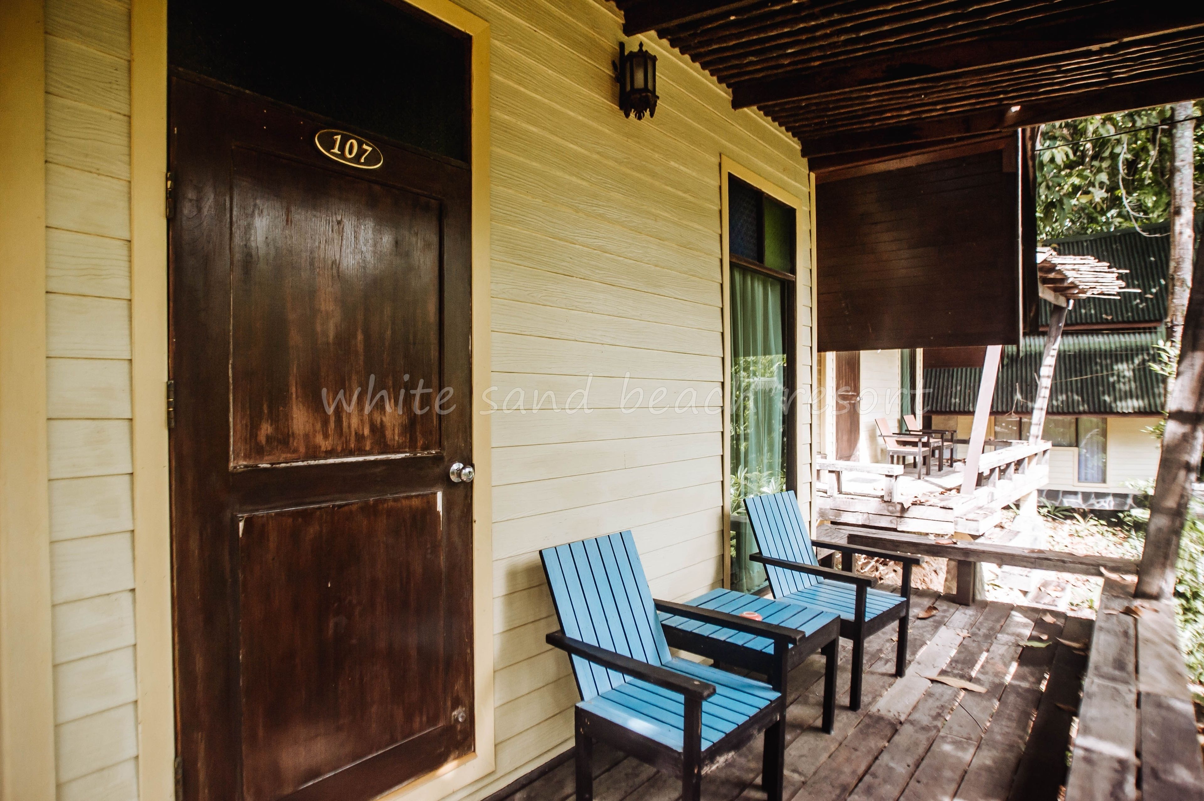 Een houten deur met een nummerbord, een veranda met twee blauwe stoelen en een overdekt zitgedeelte buiten.