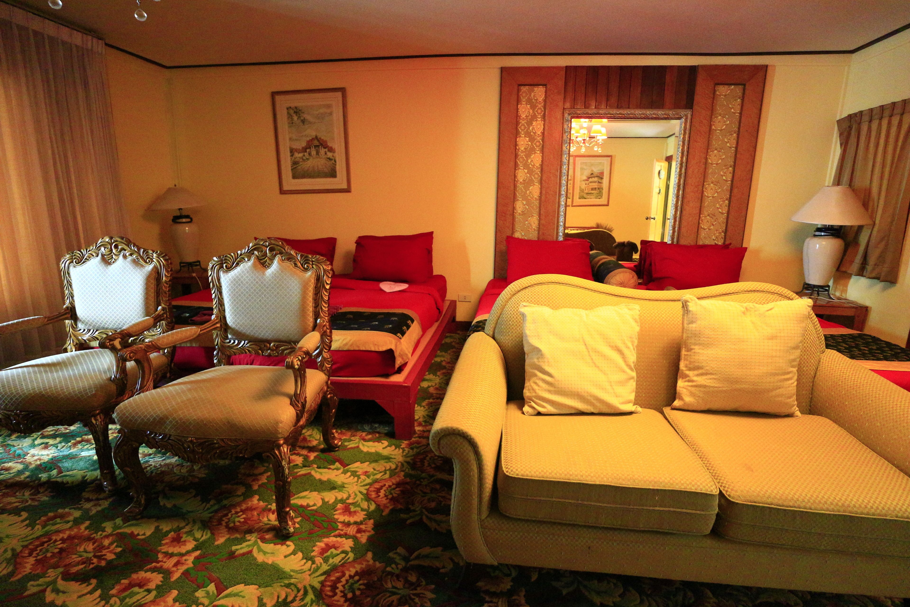 Habitación de hotel con alfombra floral, una cama roja, un sofá color beige y sillas de madera ornamentadas.