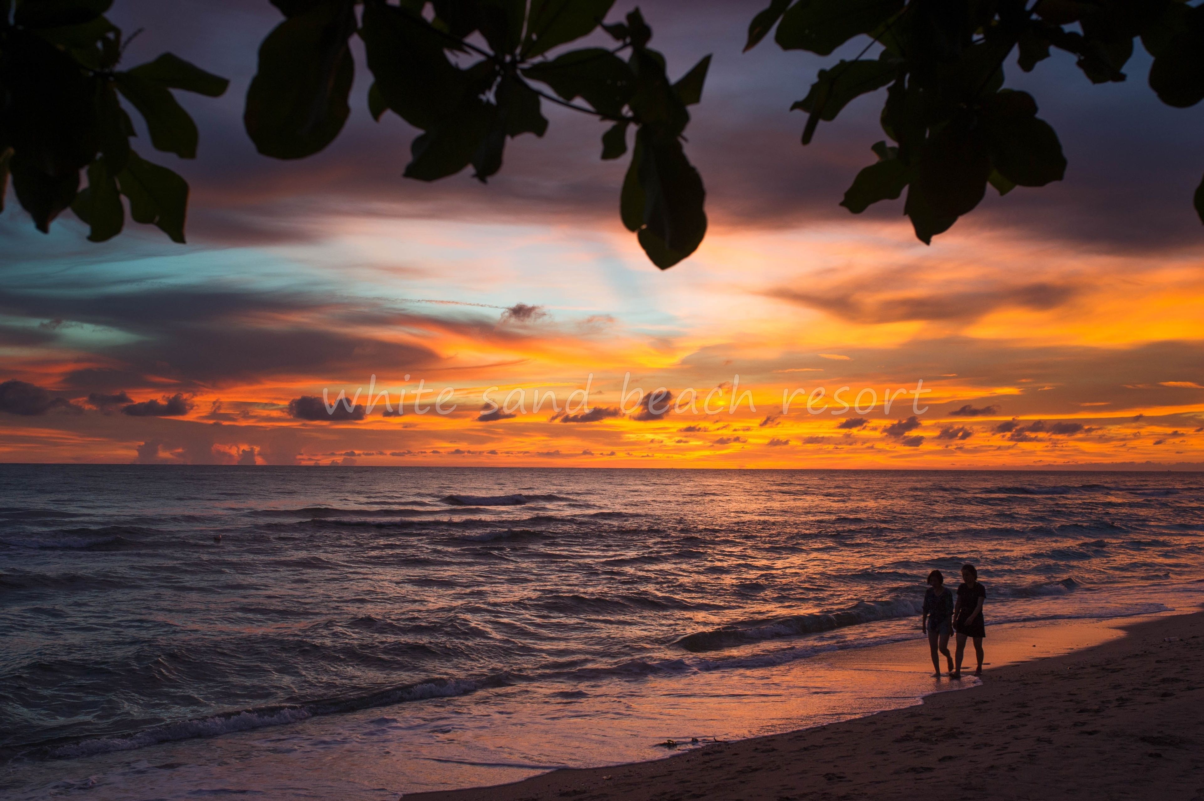 Escena de playa con dos personas caminando por la orilla al atardecer.