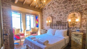 A bedroom with a stone wall, a bed with white linens, a chandelier, and a nightstand with a lamp. - Ziyabey Konagi (Ayvalik)