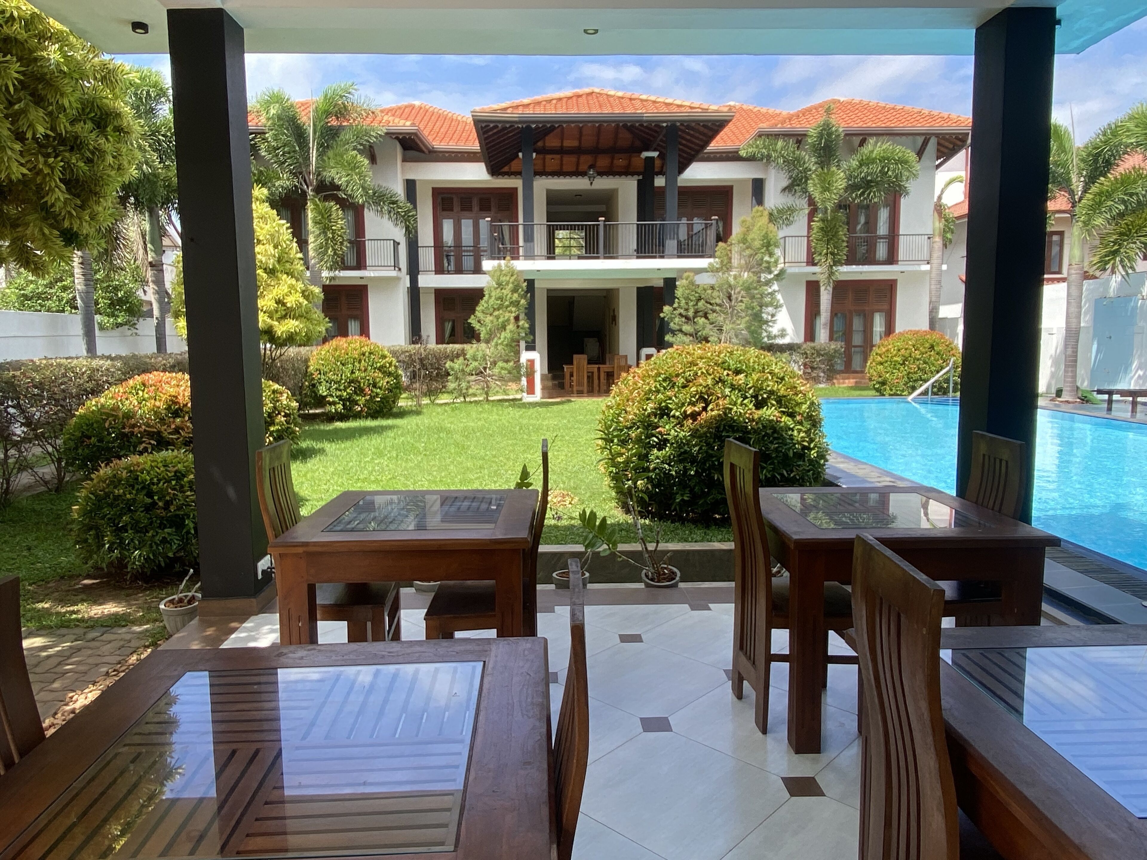 A covered outdoor area with tables and chairs, a swimming pool, and a building with a tiled roof in the background.