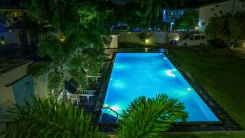 A rectangular swimming pool with a tiled deck, surrounded by lush greenery and palm trees.