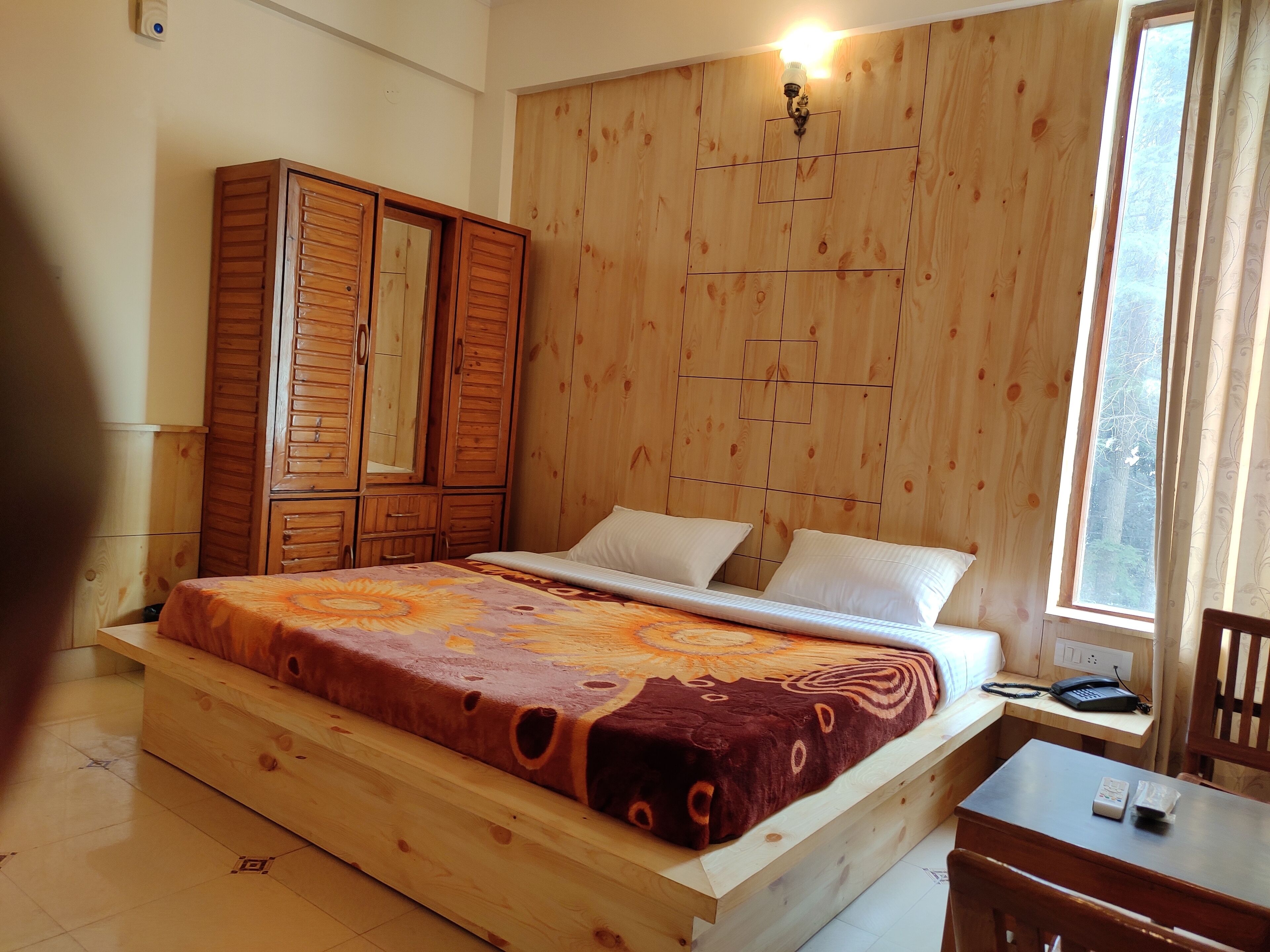 A wooden bed with a patterned bedspread, a wooden wardrobe, and a desk with a telephone and remote control.