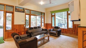 A spacious living room with wooden walls and large windows, furnished with a sofa and chairs. - Greenwoods Inn (Dharamshala)