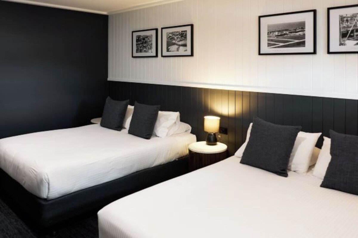 Habitación de hotel con dos camas, una mesita de noche con lámpara y tres cuadros en la pared.