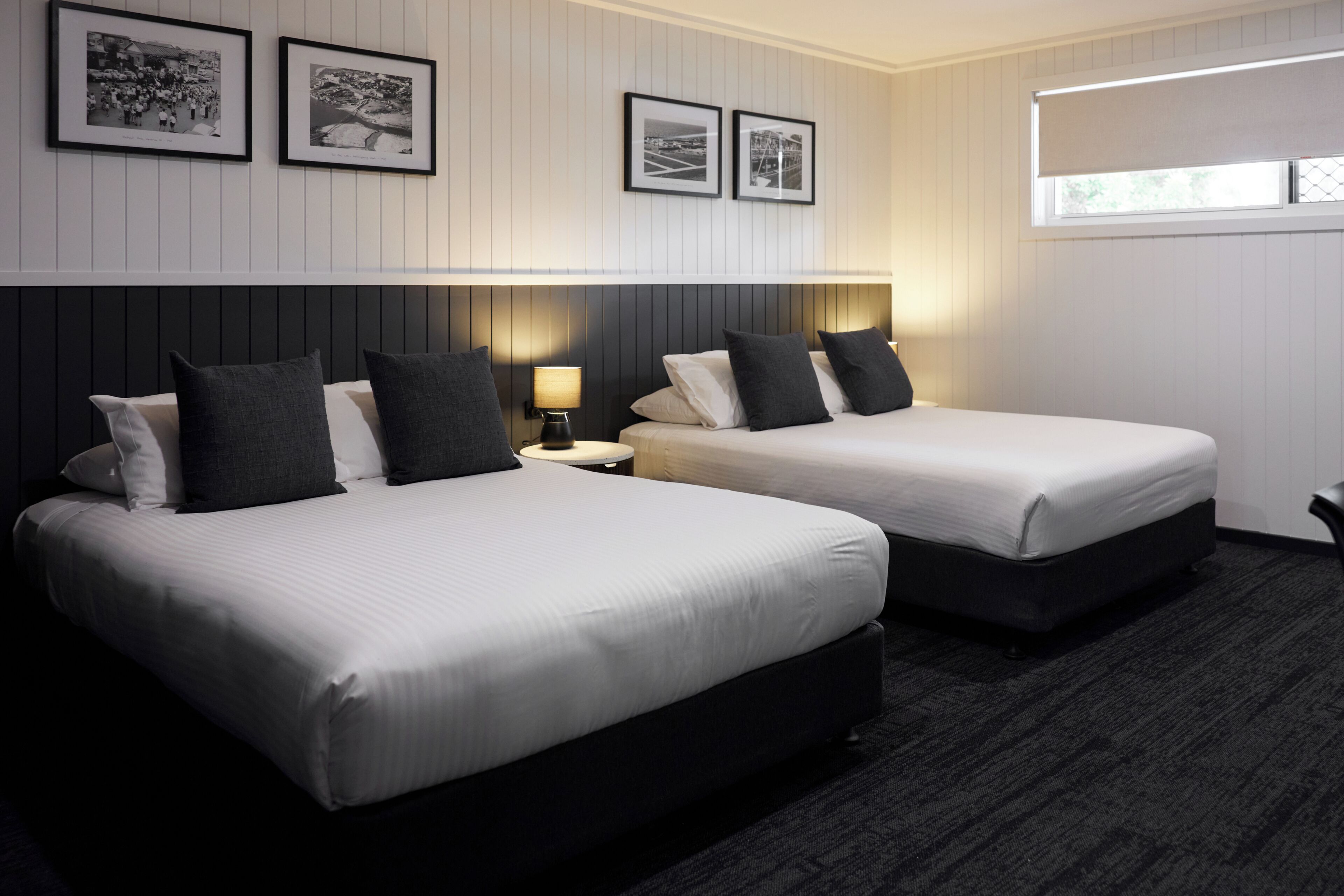 A hotel room with two beds, a nightstand with a lamp, framed pictures on the wall, and a window with blinds.