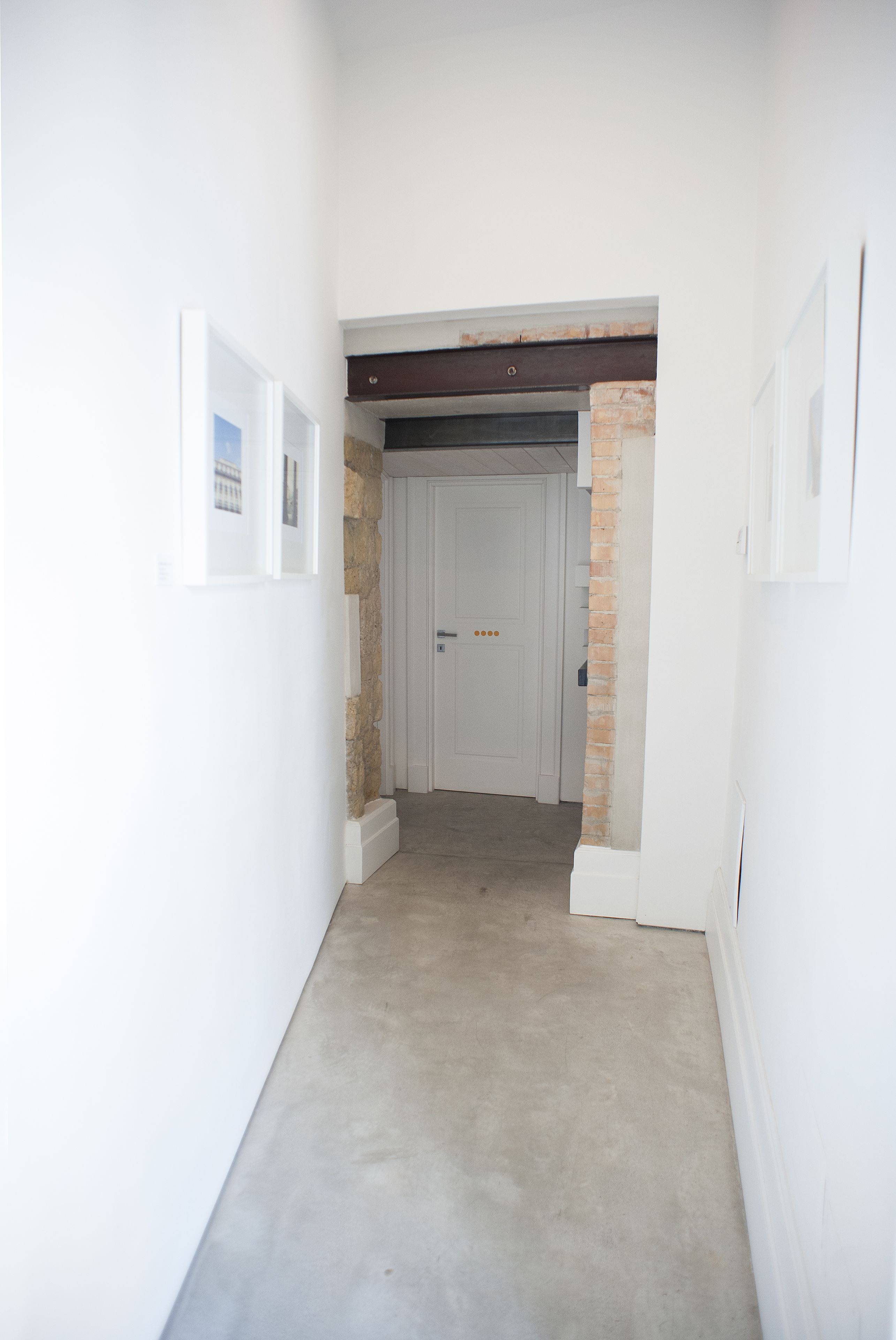 Un couloir étroit aux murs blancs, avec une porte au fond et un mur en briques apparentes.