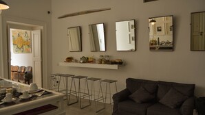 Una sala de estar con un sofá gris, una mesa de centro y una zona de bar con taburetes. Hay varios espejos en la pared y un estante con objetos decorativos.