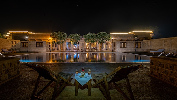 Vista nocturna de una zona de piscina con paredes de piedra, sillones de descanso y una mesa para dos con vasos y un postre.