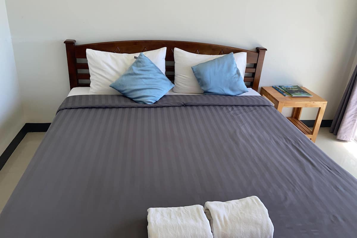 A neatly made bed with a dark grey bedspread and two pillows, one white and one blue. There is a wooden nightstand with a book on it and a wooden table beside the bed.