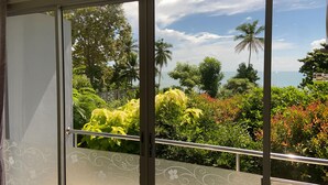 A view through a glass door with a metal railing, leading to a balcony with lush greenery and a clear sky.